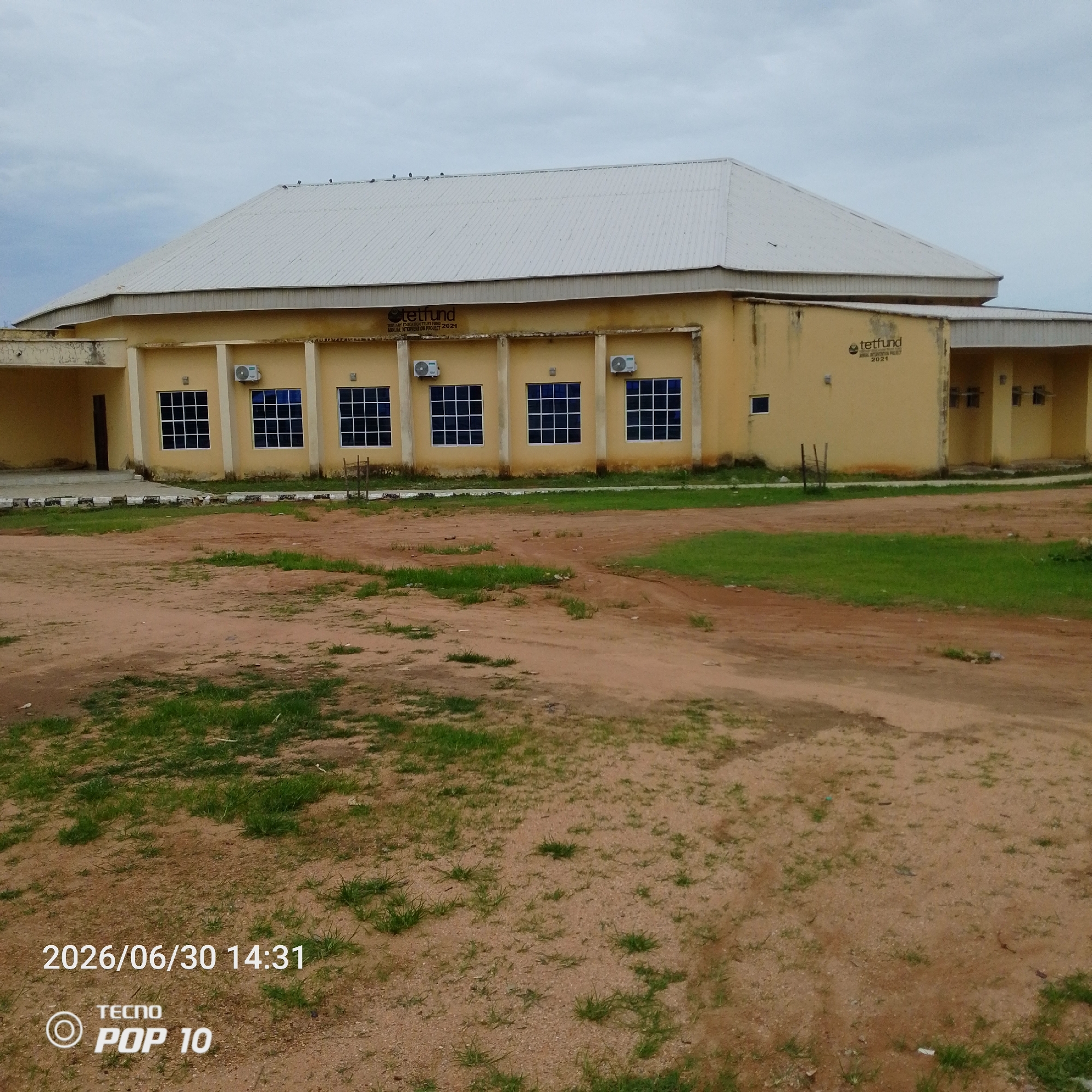 This media file shows a building with a light-colored, corrugated metal roof and yellow walls. The building appears to be a single-story structure with several windows. Some of the windows are dark blue and have a grid pattern, while others are smaller and lighter in color. Air conditioning units are visible mounted on the exterior walls.
In front of the building is an open, unpaved area with patches of dry dirt and sparse green grass. The ground appears to be a mixture of sand and small stones. The sky is overcast with clouds, suggesting a cloudy or possibly rainy day.
Visible text on the building indicates "TETFUND ANNUAL INTERVENTION PROJECT 2021". In the lower left corner of the image, there is also a timestamp "2026/06/30 14:31" and text identifying the phone used to capture the image: "TECNO POP 10".
The scene depicts an exterior view of an institutional building, likely in an educational or administrative setting, given the "TETFUND" signage. The time is mid-afternoon. There are no people visible in the image, and the overall impression is one of an unpopulated, possibly utilitarian structure. The environment appears somewhat arid, with the grass struggling to grow in the sandy soil.