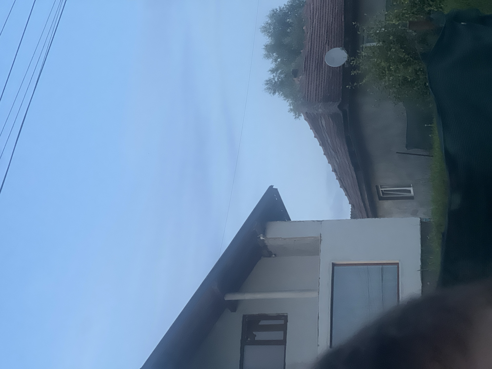 The image depicts two houses against a light blue sky with visible power lines. The house in the foreground on the left is partially under construction, with white walls and a dark-tiled roof. A large window with a dark frame is visible on its lower level. To the right, a smaller, older house with a terracotta-tiled roof and grey walls is seen. This house has a satellite dish mounted on its roof and a small window. Green bushes and grass surround the houses. The scene is an outdoor setting in Bârnova, Romania.