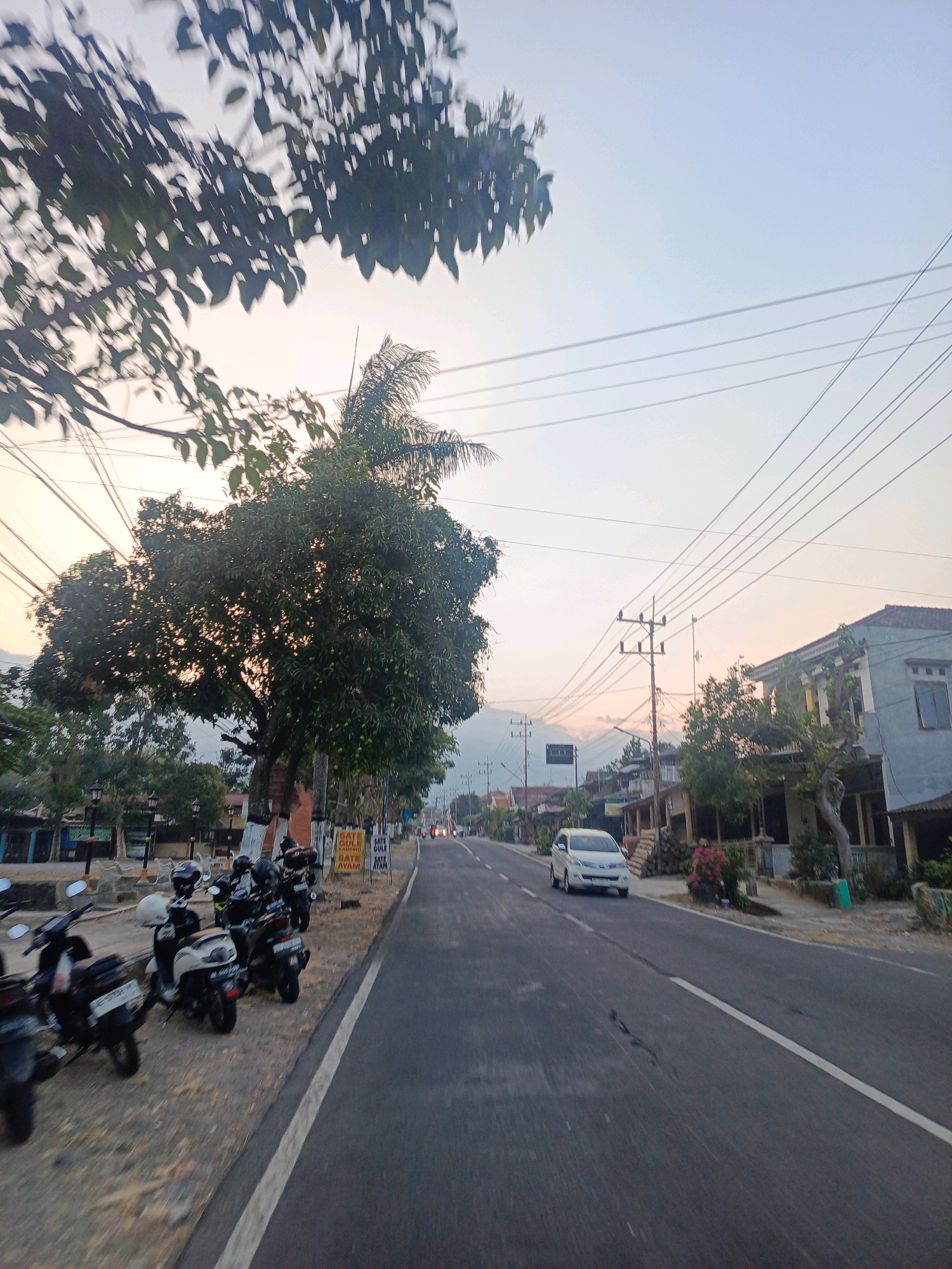 This image captures a street scene in the city of Kalang, Indonesia, during what appears to be late afternoon or early evening. The perspective is from a moving vehicle, looking down a paved road that curves slightly to the right.
On the left side of the road, a row of parked scooters is visible. These scooters are primarily black and white, some with helmets on the handlebars. A large, leafy tree with green foliage dominates the left-middle ground, its branches extending over the road and the parked scooters. Behind the scooters and the large tree, there are some buildings, including a structure with a blue facade.
The road itself is asphalt, marked with a white line down the center. A single white car is traveling on the road in the middle distance, heading away from the viewer. Further down the road, more buildings are visible, some with red-tiled roofs, and a few other vehicles, including motorcycles, can be seen. Power lines run across the sky, supported by wooden poles.
The sky is a pale, hazy blue, transitioning to a soft orange or pink hue near the horizon, suggesting either sunrise or sunset. The overall lighting is subdued, characteristic of twilight. The environment appears to be a mix of residential and commercial areas, with trees and vegetation interspersed with buildings. The atmosphere seems peaceful and calm.