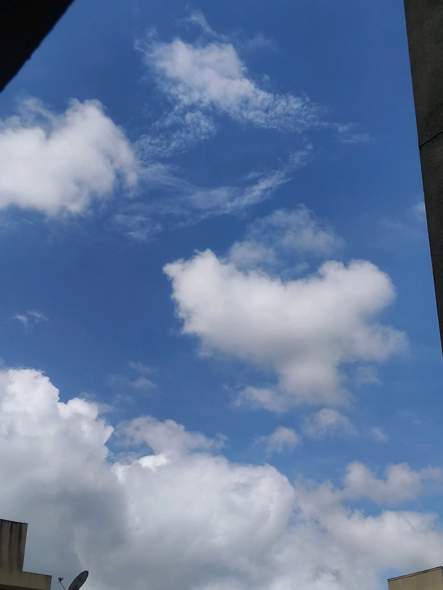 A vertical, slightly low-angle outdoor shot captures a blue sky with scattered white clouds. In the foreground, the top portion of a building is visible on the left side, with a satellite dish mounted on its exterior. The right side of the frame is partially occupied by a dark, vertical structure, likely another building. The scene is consistent with an urban environment in Pimpri-Chinchwad, India, during daylight.