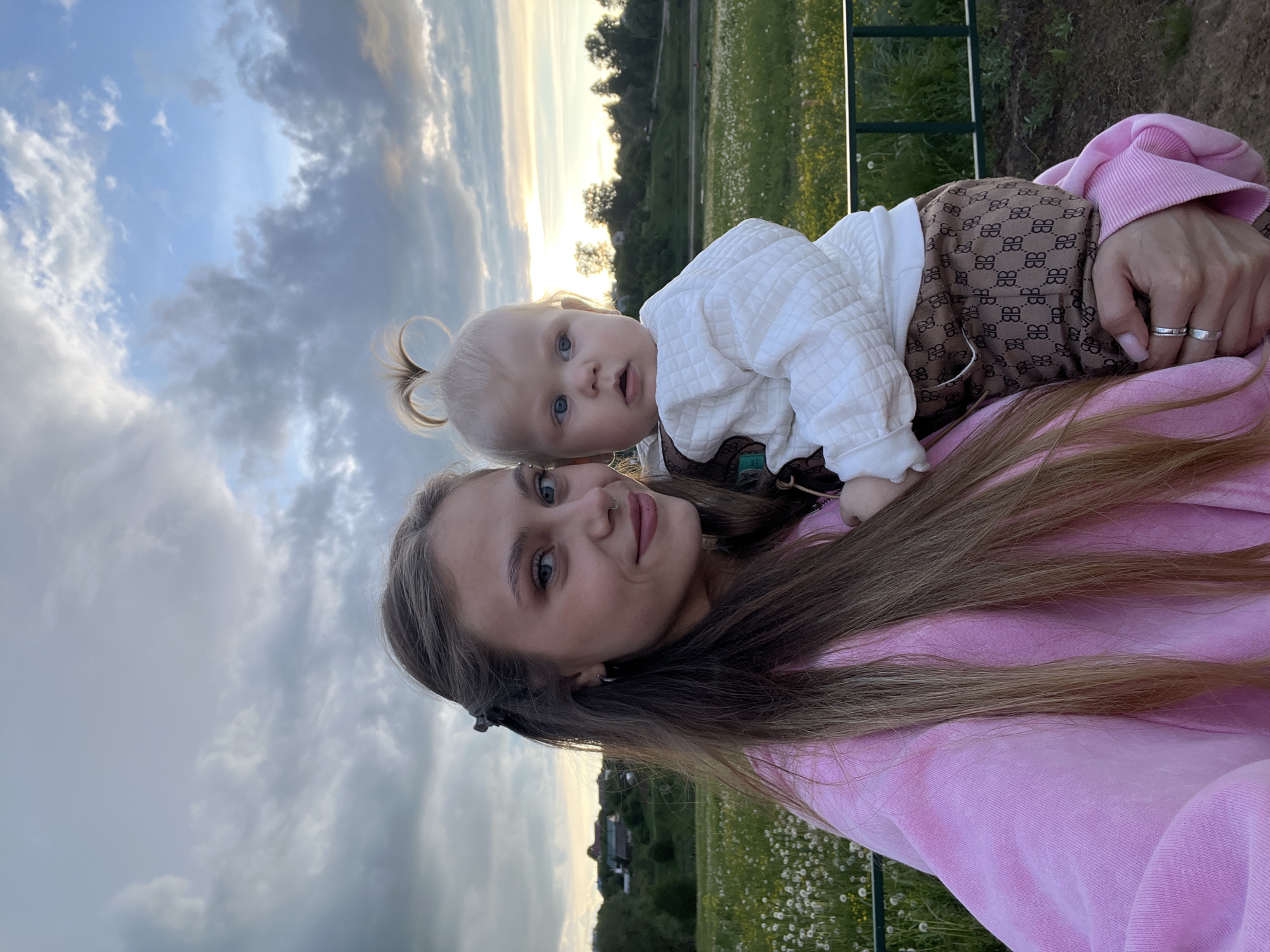 A woman in a pink sweatshirt holds a baby in brown overalls against a backdrop of a grassy field filled with dandelions, under a sky with dramatic clouds and a setting sun. The location is near Фоки, Russia, identifiable by the presence of rural architecture and dandelion-filled fields characteristic of the region. The woman looks at the camera. The baby looks at the camera with an open mouth.