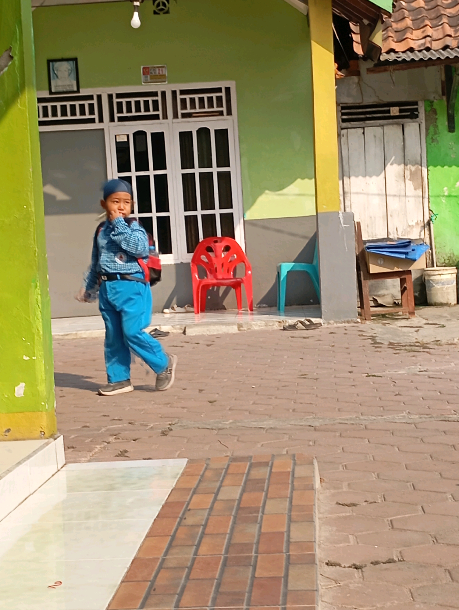 A young boy wearing a blue cap, a checkered blue long-sleeved shirt, blue pants, and dark shoes is walking on a brick pathway in front of a light green building. He has a backpack with red accents on his back and is holding his right hand to his mouth. To his left, a pillar is painted bright green and yellow. Behind him, there is a white-framed door with glass panes. A red plastic chair is to the right of the door, and a light blue plastic chair is further to the right. A wooden chair with blue items on it stands to the right of the blue chair. The building has a light green facade with a yellow pillar supporting an overhang. The ground is paved with hexagonal bricks, and there is a white tiled area to the left. The image appears to be taken outdoors in an urban or semi-urban environment.