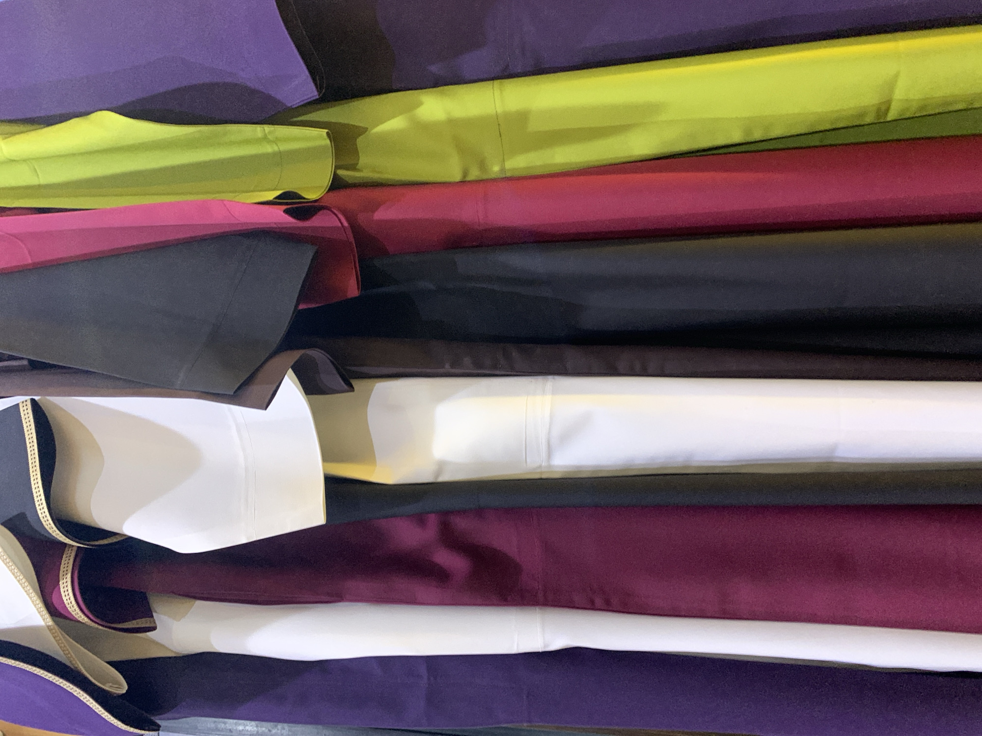 The image shows a close-up, waist-up view of several garments hanging in a row. The garments are of various colors, including purple, white, burgundy, black, fuchsia, and lime green. They appear to be made of smooth fabric and have a clean, finished look. The lighting suggests an indoor setting, possibly a shop or a storage area. There are no people or actions depicted in the image. The location in the city of Gura Top, Nigeria, is not discernible from the visual information provided.