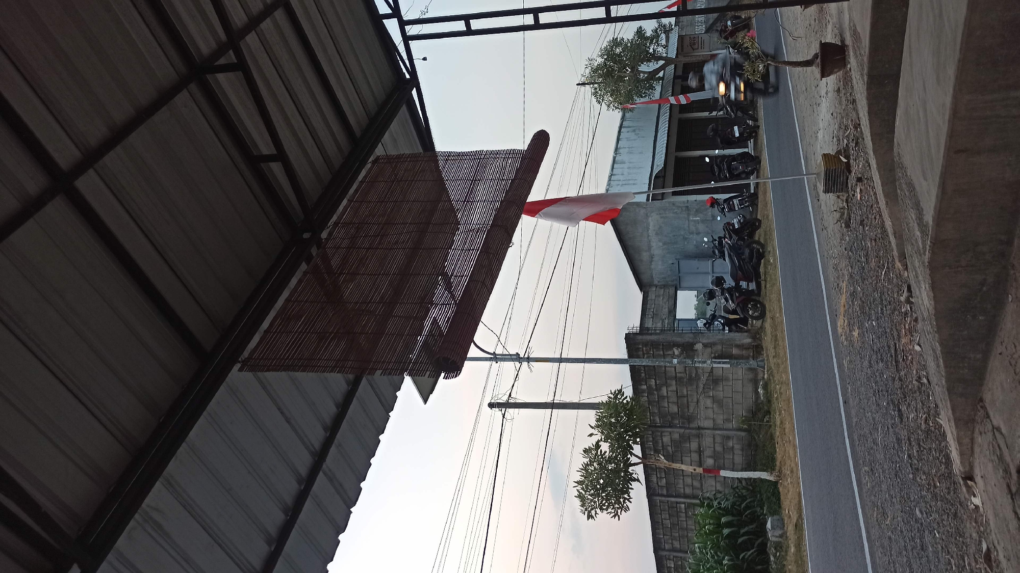 The image captures a roadside scene in Bedikulon, Indonesia, likely during the late afternoon or early evening due to the soft, diffused light and the colors of the sky. The foreground is dominated by a concrete structure, possibly a bench or platform, with visible gravel and debris scattered around.
A paved road runs horizontally across the mid-ground, with a white dashed line indicating the lane. On the left side of the road, there is a grey brick wall. In front of the wall, a young tree with a white and red striped trunk stands. To the right of the tree, several motorcycles are parked along the roadside. Further to the right, a building with a grey facade and a metal awning stretches across the scene. A tall pole with a bamboo blind hanging from a structure above the road is positioned near the building.
A prominent Indonesian flag, red and white, is flying from a pole in front of the building. A person on a motorcycle is in motion on the road, creating a slight blur. In the background, there are more power lines stretching across the sky, and the horizon suggests open land or fields beyond the structures. The overall impression is a simple, functional outdoor space adjacent to a road, common in many Indonesian towns.