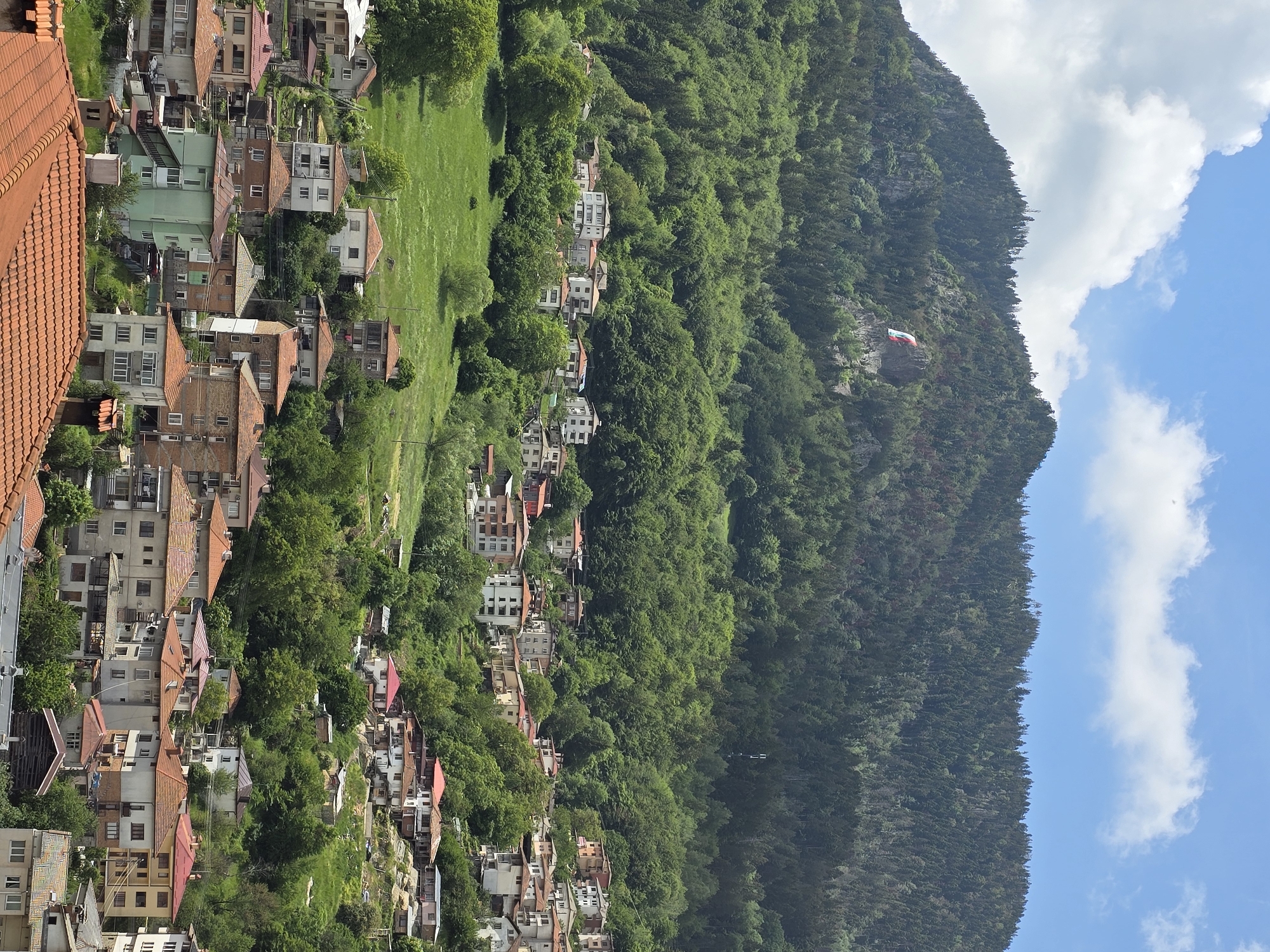 The image presents an expansive view of a mountain village or town set against a backdrop of a densely forested mountain and a clear blue sky. The original image is rotated 90 degrees clockwise.
In the foreground, a close-up of a tiled roof, predominantly terracotta in color, is visible, suggesting the photograph was taken from an elevated position. Below this, a sprawling settlement extends across the hillside, characterized by numerous multi-story houses. Most buildings feature traditional orange or red tiled roofs, while some have flatter, grey, or brown roofs. The houses exhibit varied architecture, often with white or light-colored facades, and are interspersed with abundant green trees and patches of grassy terrain.
The village gradually transitions into the natural landscape, with more houses built into the lower slopes of a large, steep mountain. This mountain, heavily covered in dark green coniferous and deciduous trees, dominates the midground and background. Prominently displayed on a rocky outcrop or clearing on the mountainside is a large banner or flag, characterized by horizontal stripes of red, white, and green, indicative of the Bulgarian flag.
Above the mountain, the sky is a bright, clear blue, adorned with several fluffy white clouds. The scene is bathed in bright daylight, suggesting a sunny late morning or afternoon. No people or specific activities are visible, and there is no discernible text in the image. The overall impression is of a scenic, mountainous region, likely in Eastern Europe, on a pleasant day.
