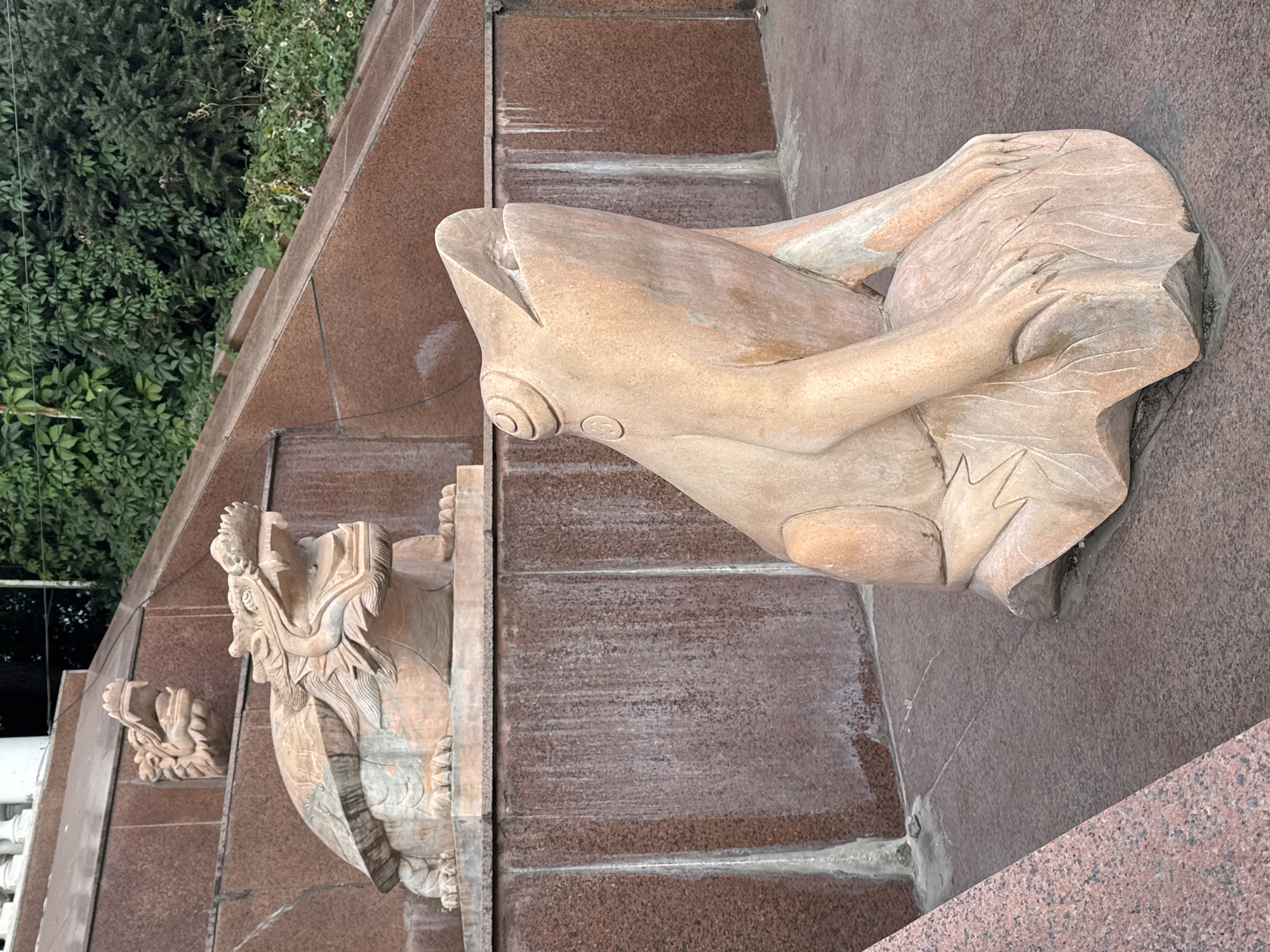 In Koy Tash, Kyrgyzstan, a stone sculpture of a frog sits on a rock base in the foreground, facing right. The frog sculpture is a pale pinkish-tan color and appears weathered. Behind the frog, a tiered structure made of reddish-brown granite with lighter streaks rises. On the second tier of this structure, a stone sculpture of a dragon turtle is visible. The dragon turtle is also a pale pinkish-tan color and is depicted with its mouth open, revealing teeth. Its body is carved with details resembling scales and a shell. Above the dragon turtle sculpture, a smaller, carved dragon head protrudes from the granite wall. Lush green foliage, including trees and vines, forms a dense backdrop behind the granite structure. The ground in front of the frog sculpture is paved with reddish-brown tiles. Water stains or streaks are visible on the granite surfaces.