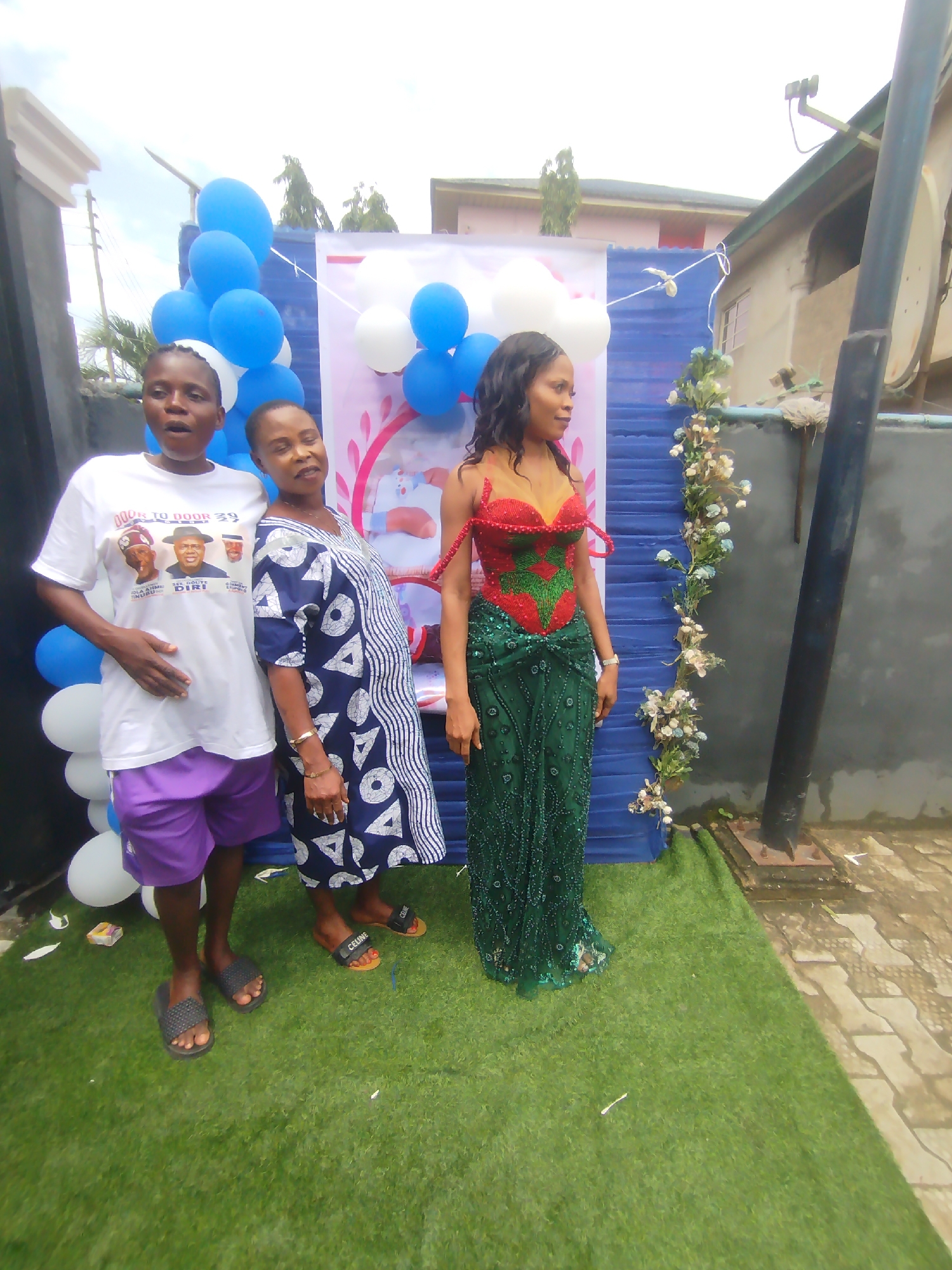 In Yenegoa, Nigeria, three women pose for a photo in an outdoor setting, possibly a yard or event space. The woman on the left wears a white t-shirt with a graphic of three men, purple shorts, and black slides. The woman in the middle wears a blue and white patterned dress and black sandals. The woman on the right is dressed in a sparkling green beaded gown with a red and green beaded bodice, and she stands to the right of the other two. Behind the women is a blue and white banner decorated with balloons. To the right of the banner, a staircase is adorned with a floral garland. The ground is covered with artificial grass and pavers. A dark pole stands to the right of the scene.