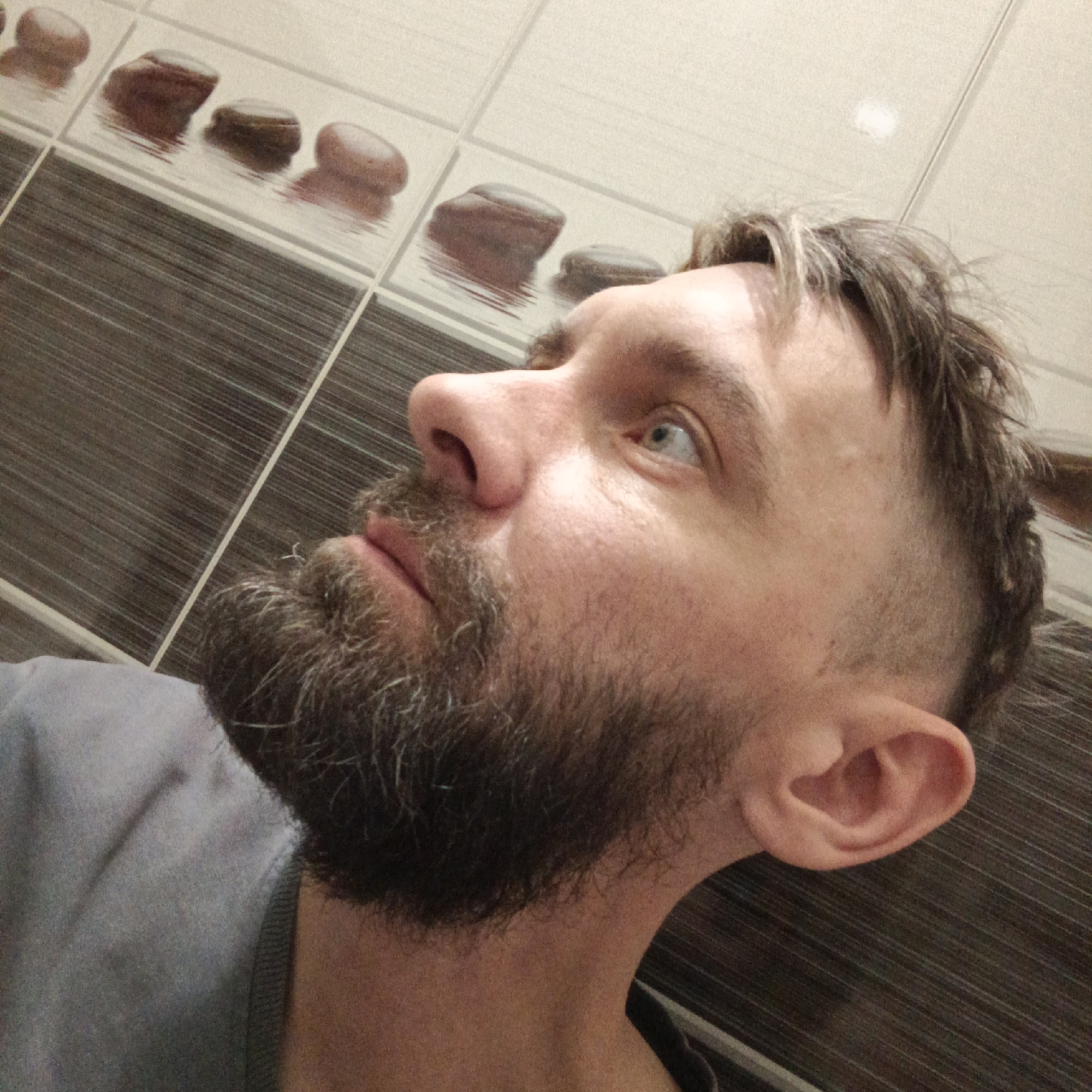 A middle-aged man with a beard is looking upwards. He has light eyes and short, graying hair. He is wearing a gray shirt. The background appears to be a tiled wall with a horizontal stripe pattern and decorative stone images. The location is likely a bathroom. There is no indication that this image was taken in or near the Russian city of Марковский.