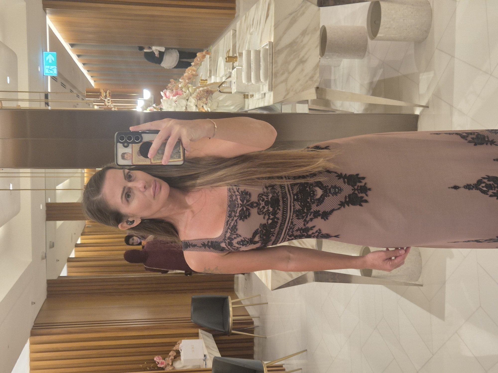 A woman takes a mirror selfie in what appears to be a luxurious indoor setting. She is standing upright, holding a black smartphone with a decorative case in her right hand. Her long, straight brown hair flows over her shoulders, and she wears a light brown or mauve dress with intricate black lace patterns on the bodice and sides. A small earpiece is visible in her left ear, and a gold bracelet adorns her right wrist.
The background reflected in the mirror reveals a refined space. To her right, a bright green exit sign with a left-pointing arrow is seen above wooden slatted walls. Below this, a display features light-colored flowers, including pink and white blossoms, arranged on a marble surface. A person in a black and white uniform, possibly staff, is faintly visible in the background.
To her left, the reflection shows a high-end vanity or bathroom area. A marble counter is equipped with gold-colored fixtures and three wall-mounted dispensers, one of which is partially labeled "shampoo." Further back, two large, light-colored cylindrical objects are present. The floor is tiled with a light, geometric pattern. In the lower foreground, two dark chairs with gold frames are visible, along with a white box that may be a luxury item, possibly bearing the brand "Dior," and additional floral arrangements.