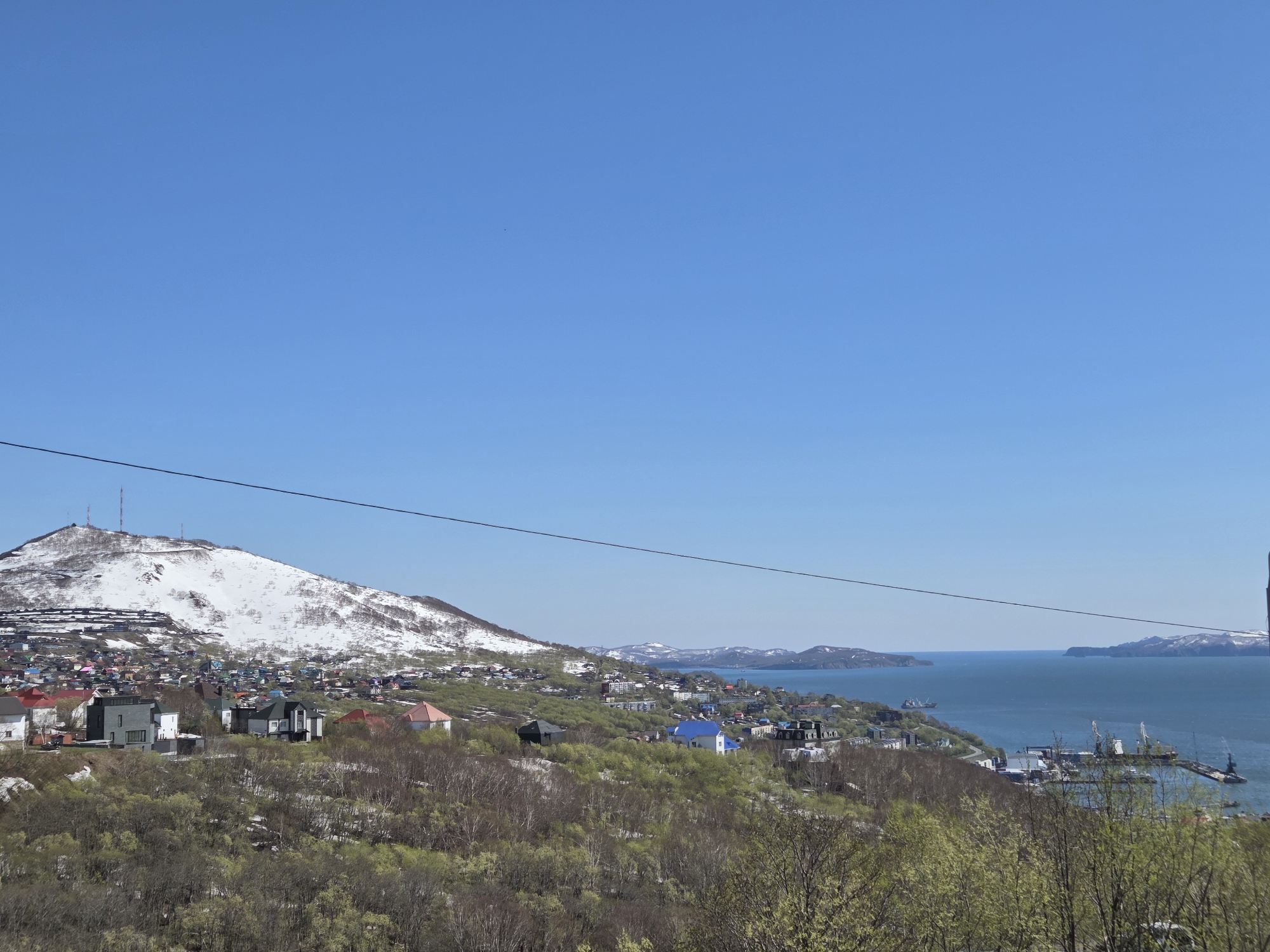 The image captures a panoramic view of a coastal city nestled amongst hills and a body of water, potentially a bay or sea, under a clear blue sky. A snow-capped mountain occupies part of the left side of the image, with antenna or communication towers on its peak. Buildings and houses of varying colors and styles are scattered across the lower slopes of the mountain and along the coastline. Patches of green vegetation dot the landscape. On the right side of the image, a port area with cranes and what appears to be a ship is visible near the water's edge. A power line stretches across the frame, intersecting the scene horizontally. The overall composition suggests a settled, possibly remote, location.