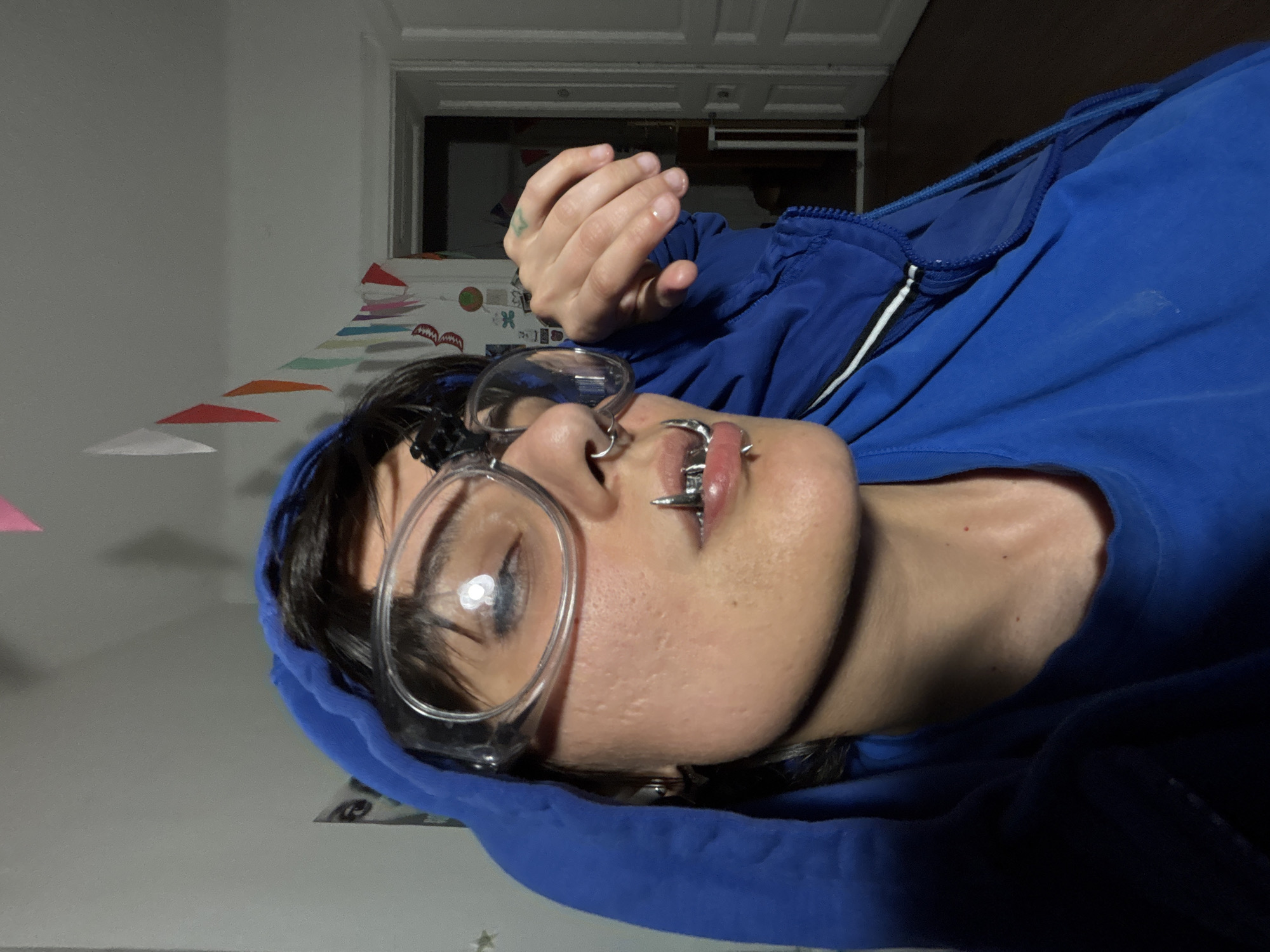 A person with dark hair and glasses is lying down, wearing a blue hooded sweatshirt. They have lip piercings. A hand is raised near their face. Behind them is a wall with colorful triangular decorations and a doorway. The location is unknown and cannot be determined from the image.