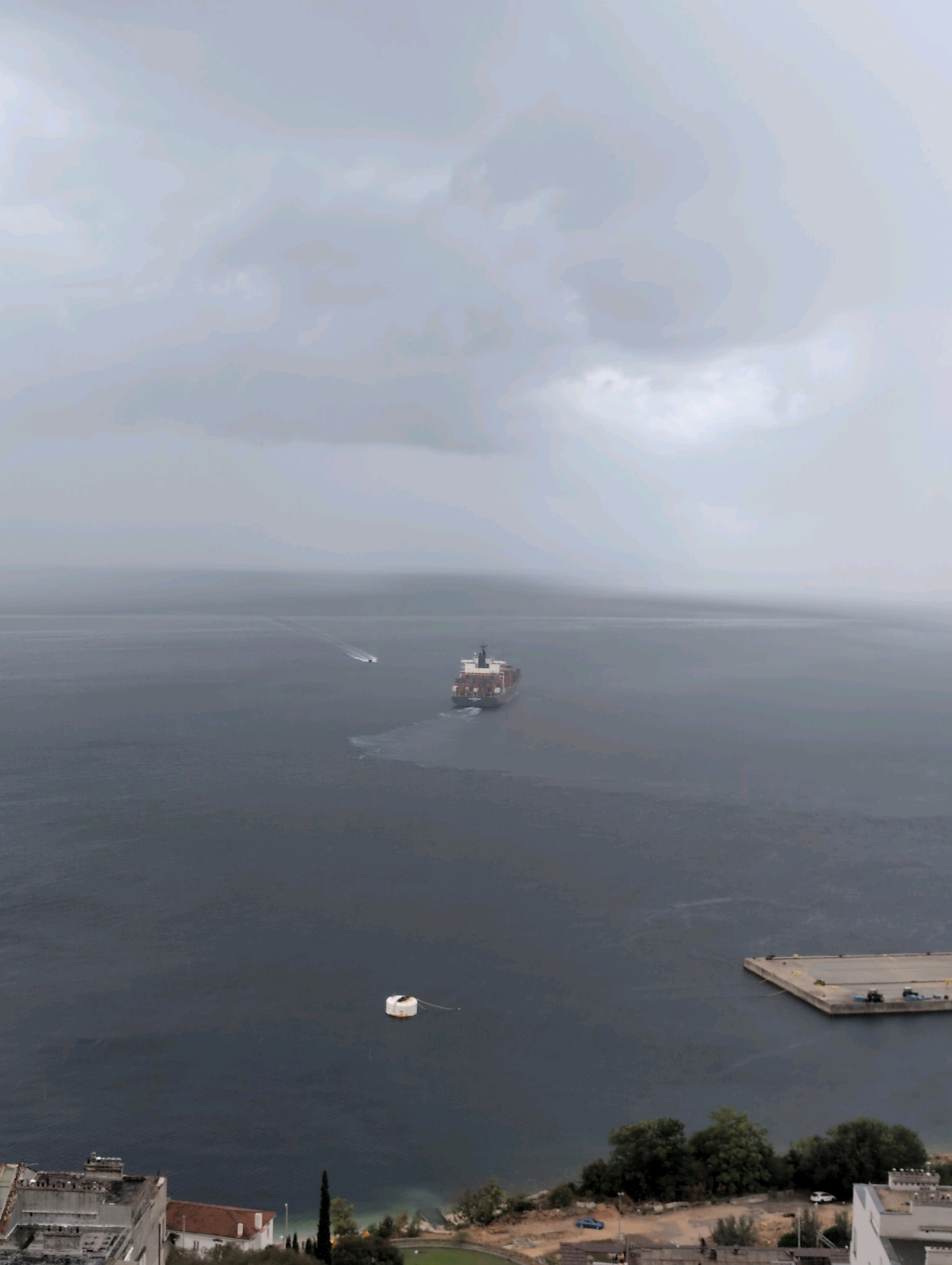 The aerial image, taken in Grad Rijeka, Croatia, captures a vast expanse of a calm, dark sea under a heavily overcast sky. In the distance, a large cargo ship is visible, leaving a white wake as it moves through the water. A smaller boat is also present, further ahead and to the left of the cargo ship. Closer to the foreground, a solitary white buoy floats on the surface of the water, tethered by a rope. The coastline is lined with buildings and trees. To the right, a concrete pier extends into the sea, with what appear to be vehicles or equipment on it. The overall atmosphere is muted and somewhat somber due to the overcast conditions.