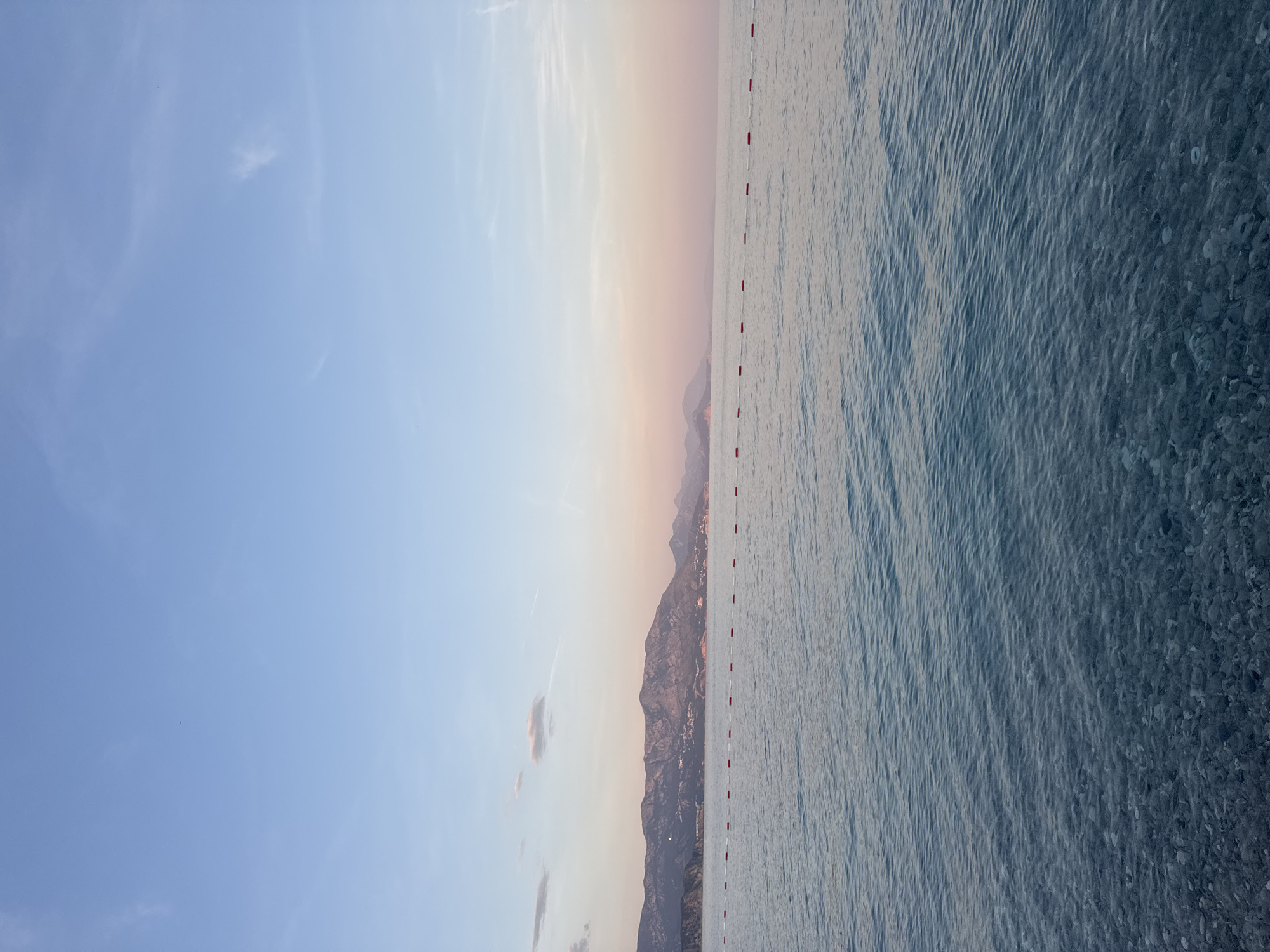 The image shows a beach scene in Prijevor, Montenegro. The sky transitions from light blue to a pale orange near the horizon. The sea is a light blue color and appears relatively calm, with the foreground showing more texture and depth indicating shallow water over pebbles. A mountain range is visible along the horizon. A string of red and white buoys runs vertically along the edge of the water.