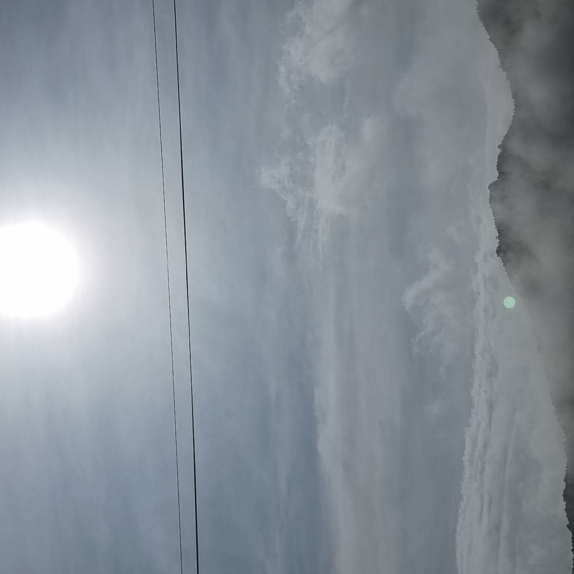 The image depicts a hazy, overcast sky with the sun shining brightly at the top center, creating a diffused glow. Two dark, thin wires stretch horizontally across the upper portion of the frame. Below the wires, layers of clouds create a textured, muted backdrop. The lower part of the image shows the silhouette of rolling hills, partially obscured by thick fog. The hills appear to be covered in dense vegetation, indicated by dark, irregular shapes. A faint, greenish-blue circular light is visible near the center of the image, partially embedded within the fog and on the side of a hill. The overall scene suggests a natural landscape, possibly a mountain or hilly region, during daylight hours with significant atmospheric conditions like fog or haze. The location is specified as Dunnae-myeon, South Korea.