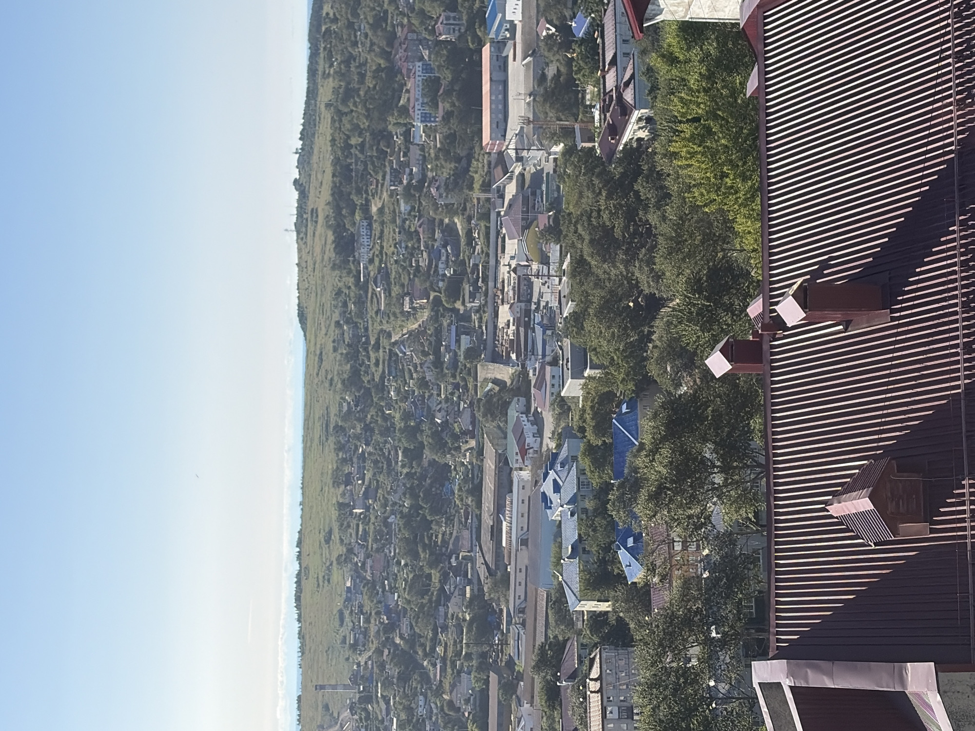 This is a high-angle, medium-shot, vertical, outdoor photograph of the city of Korsakov, Russia, on a clear day. The sky is a pale blue with a slight haze towards the horizon. The foreground is dominated by a dark red, corrugated metal roof with two small, gabled structures that appear to be roof vents or decorative dormers. The middle ground shows a dense residential area with various buildings of different sizes and colors, many featuring blue roofs. Trees are interspersed throughout the city, indicating a temperate climate. In the background, the landscape rises into a gently rolling, grassy hill dotted with more trees and some distant structures, possibly communication towers. The overall impression is of a coastal city nestled amongst rolling hills. There are no people visible in the image.