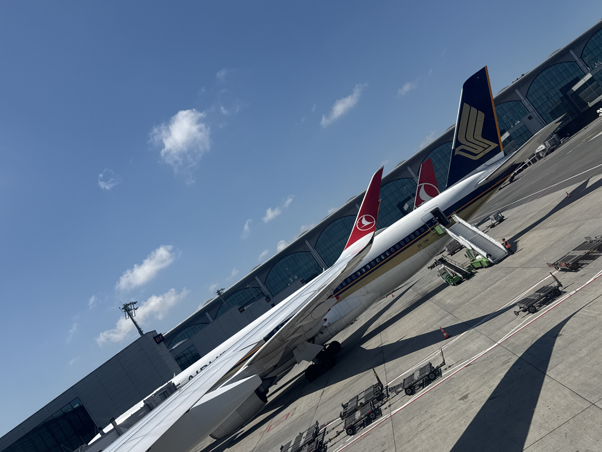 The image shows an airport scene at Arnavutköy, Turkey, under a clear blue sky with scattered white clouds. The perspective is angled, giving a sense of looking down on the tarmac. In the foreground, the wing of a large aircraft is visible, with its landing gear extended. Adjacent to this aircraft, two other airplanes are parked, one with the red and white logo of Turkish Airlines on its tail fin, and another with the dark blue and gold logo of Singapore Airlines.
Several baggage carts are lined up on the tarmac, along with a jet bridge that is connected to one of the aircraft. The airport terminal building, characterized by its modern glass and metal facade, forms the backdrop. The scene appears to be active, with ground crew and equipment present, indicating aircraft servicing operations. The lighting suggests it is daytime, likely late morning or early afternoon, given the sharp shadows cast by the aircraft and equipment.