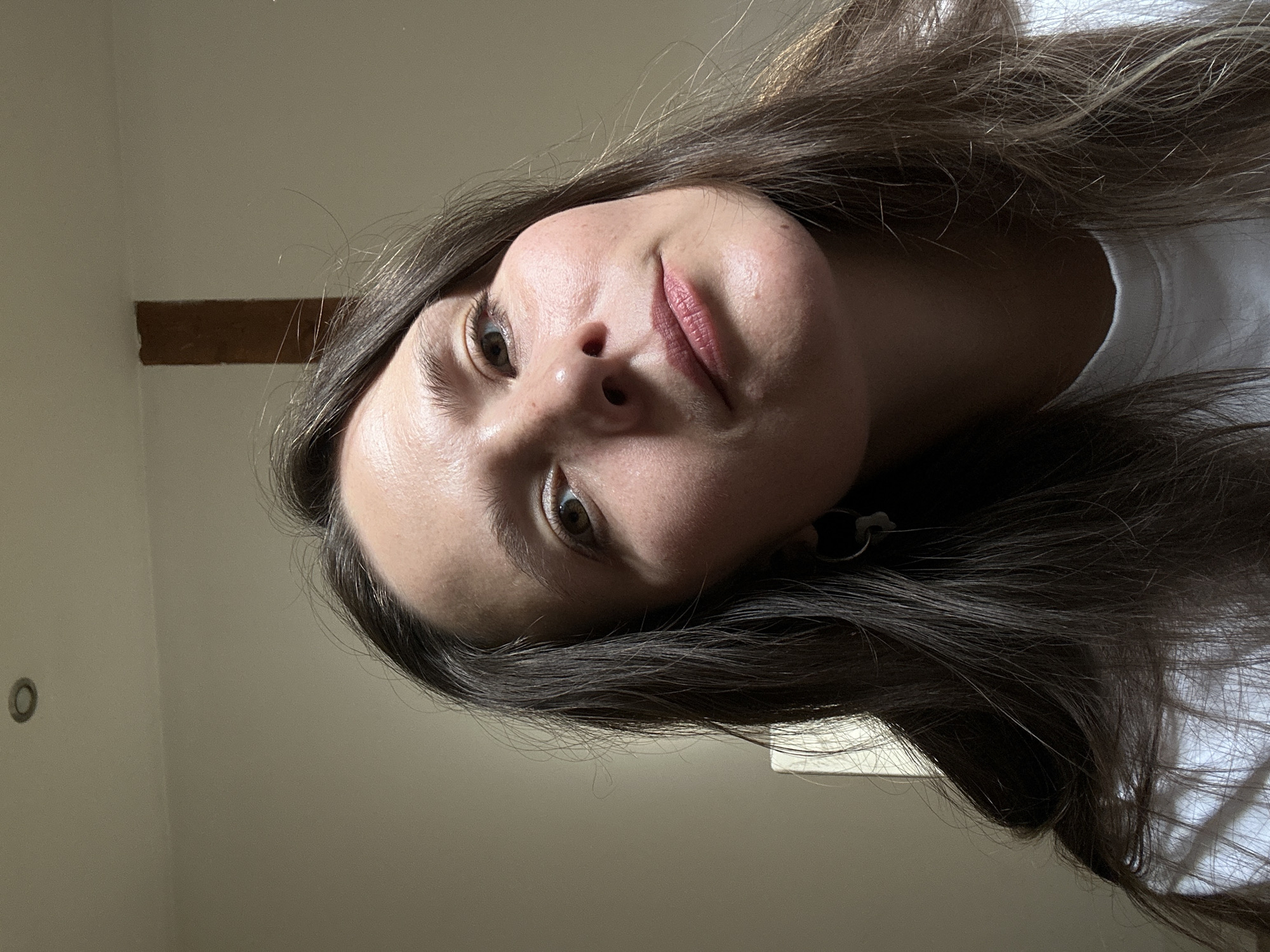 The original image, rotated 90 degrees clockwise, features a close-up portrait of a young woman. Her face, framed by long, dark brown hair, is positioned centrally and looks upwards towards the viewer with a thoughtful or slightly curious expression. She has brown eyes and light, naturally colored lips with a subtle pink tint. Her skin has a natural sheen, suggesting good lighting.
She is wearing a white top, visible on her right shoulder and upper chest area. The background suggests an indoor setting with light-colored walls. To her left (when the image is conceptually rotated upright), a dark brown, rectangular wooden beam is visible running horizontally across the wall, indicating rustic or traditional architecture. On the upper left wall, a small, circular, dark-colored object, possibly a light fixture or vent, can be seen. The lighting appears bright and natural, suggesting it is daytime. No visible text is present in the image. The setting could be a room in Bartula, Montenegro, given the location context.