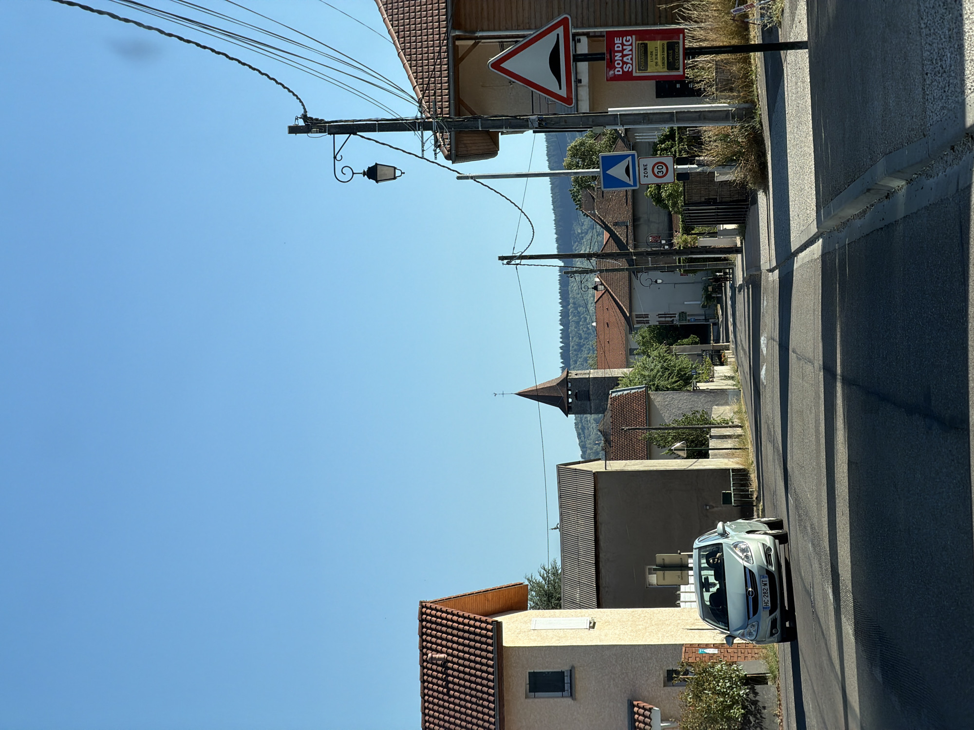 This image shows a street scene in a French village. The perspective is tilted, making it appear as though the viewer is looking down from a slight angle. The sky is a clear blue, indicating a sunny day, likely in the afternoon due to the long shadows cast across the road.
The foreground features a light-colored car parked on the right side of a paved road. The car has its headlights on and its license plate is visible, reading "HC-282-WT". A concrete curb runs along the edge of the road.
The middle ground shows several buildings with red tile roofs, typical of rural French architecture. A distinctive church tower with a pointed roof rises above the other structures. There are also street signs visible. One sign, in a triangular red border, depicts a horn. Another sign is blue with a white triangle and the word "ZONE" above a "30" indicating a 30 km/h speed limit. A red sign with white text announces "DON DE SANG" (blood donation) with dates and times.
The background is dominated by a lush green forest covering rolling hills, suggesting the village is nestled in a natural landscape. Telephone wires crisscross the sky, supported by utility poles. The overall impression is one of a quiet, sunny day in a rural French community.