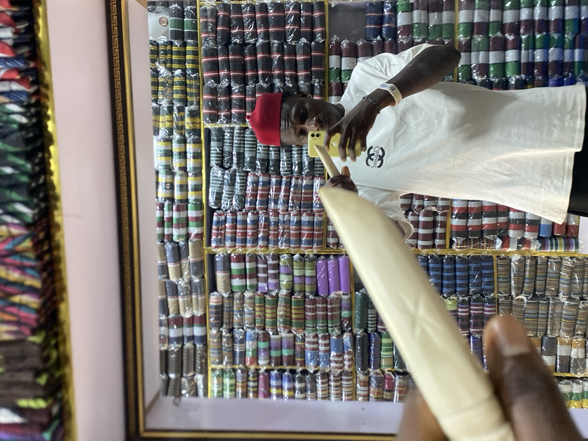 A man wearing a red cap and a white t-shirt is taking a picture with his cell phone of a display of colorful fabrics rolled up and arranged on shelves. The fabrics appear to be individually wrapped in plastic. The man's wristband is visible, and he is holding a light-colored object, possibly a pen or a stick. The scene is framed, suggesting it may be a photograph or a display in a shop. The location is identified as Bukuru, Nigeria.