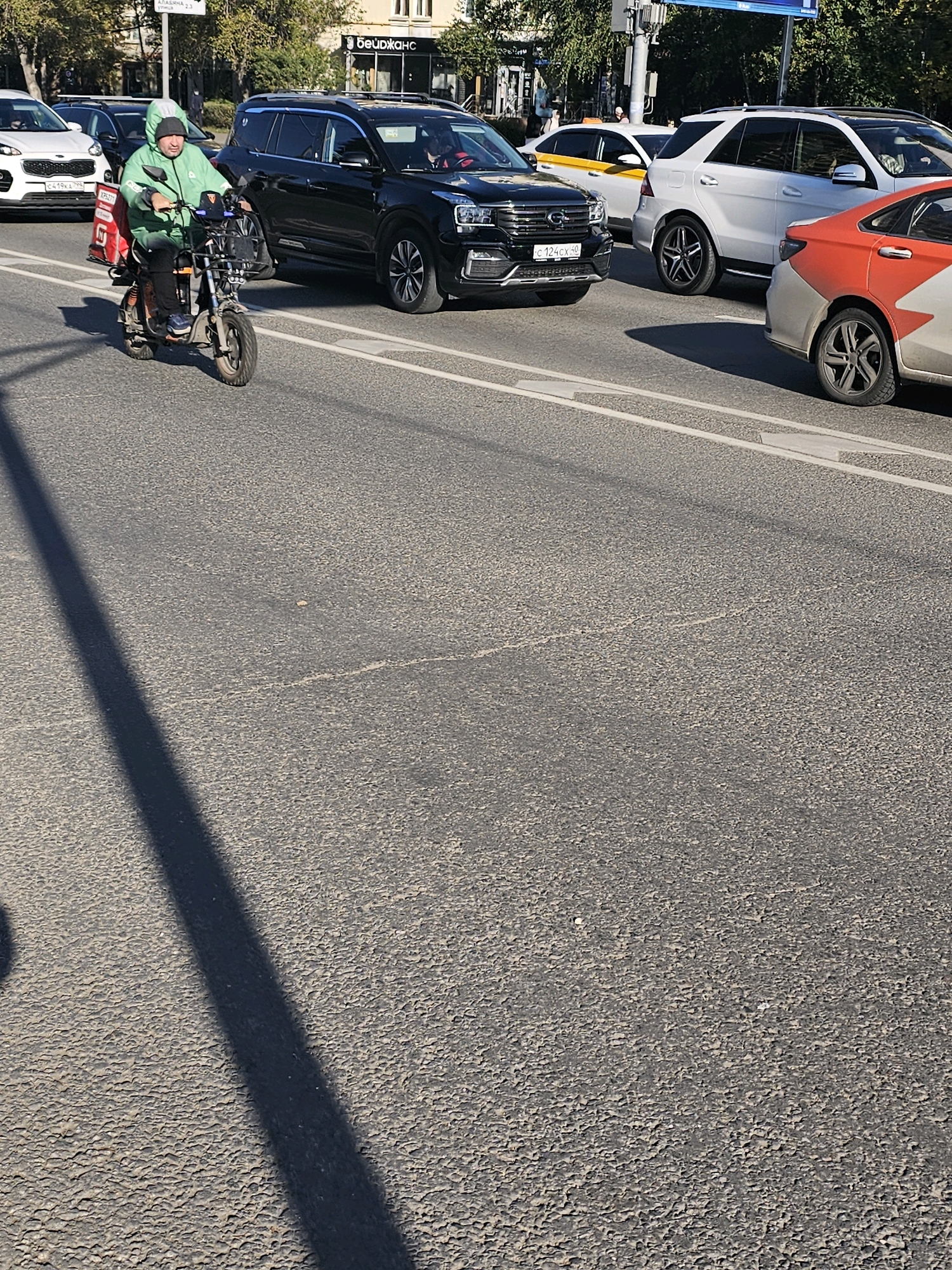 A person is riding a scooter with a large red delivery bag on the back, through a city street in Moscow, Russia. The person is wearing a green hooded jacket and a beanie. The scooter has a basket in the front. Several cars are on the road around the scooter, including a black SUV, a white SUV, a white sedan with a yellow stripe, and a red and white car. The road surface is asphalt, and there are lane markings visible. Trees and buildings are visible in the background, suggesting an urban environment. The scene appears to be captured during daylight, with shadows cast on the road.