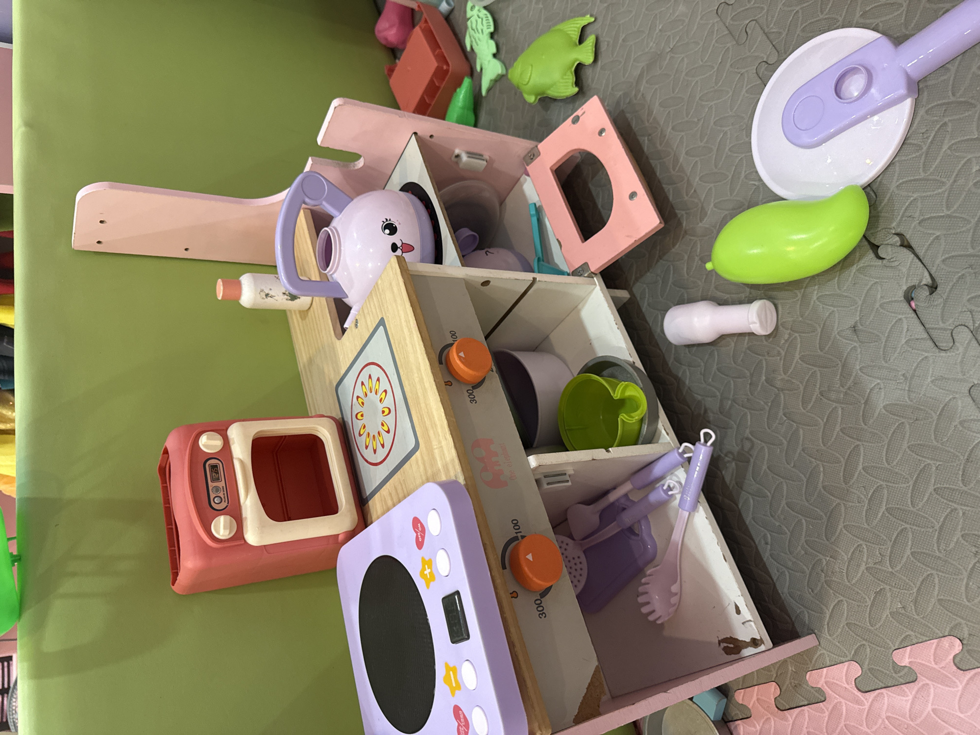 The image depicts a children's toy kitchen set, likely located in a home or daycare setting in Bac Nha Trang Ward, Vietnam. The toy kitchen is predominantly pink and light wood, featuring a pretend oven, stovetop, and various accessories. A purple kettle with a cute face sits on the stovetop, next to a white bottle with a pink cap. Below the stovetop, an open compartment reveals purple utensils and a green bowl. A pink door hangs open at the bottom of the unit. Scattered around the toy kitchen are additional play items, including a green mango-shaped toy, a purple-handled white plate with a purple utensil, and several green plastic fish. The toys are placed on a grey, interlocking foam mat. In the background, a bright green, padded wall is visible, suggesting an indoor play area. There are no people visible in the image. The overall impression is one of a well-used, but organized, play space for young children.