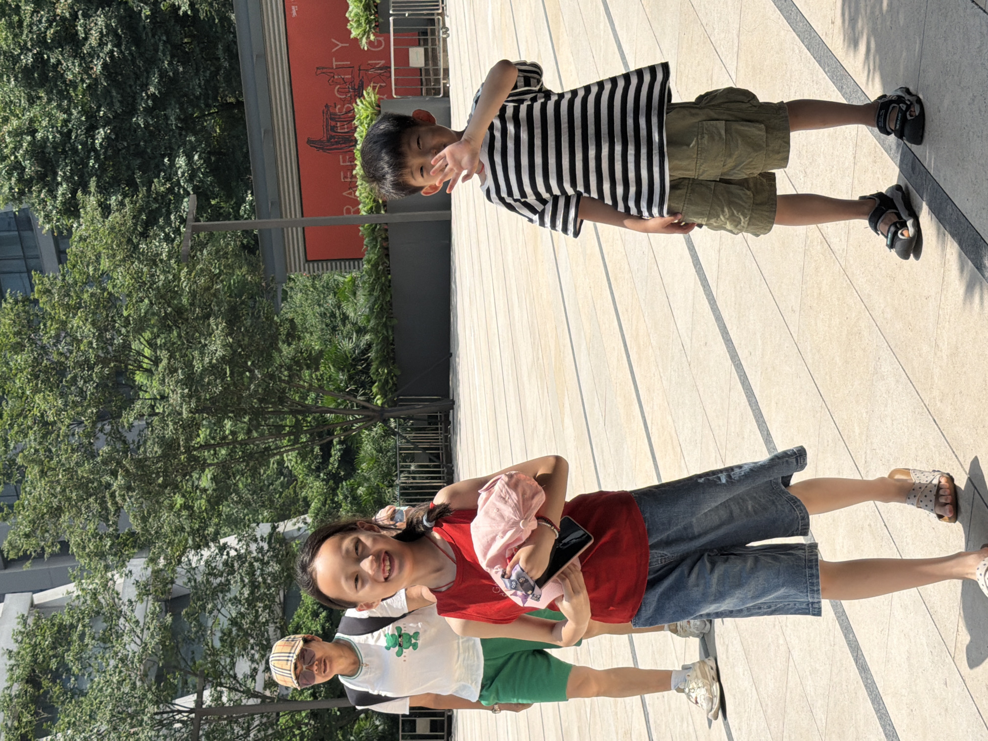In the city of Yuzhong District, China, a group of people are walking on a paved outdoor area. In the foreground, a young girl wearing a red sleeveless shirt and denim shorts is smiling at the camera, holding a pink hat and a cell phone. To her right, a young boy in a striped t-shirt and olive green cargo shorts waves to the camera. Behind the girl, an adult wearing a white t-shirt, green shorts, and a checkered hat is walking away from the camera. In the background, there are trees and a building with a red sign. The ground is paved with light-colored tiles and darker lines.
