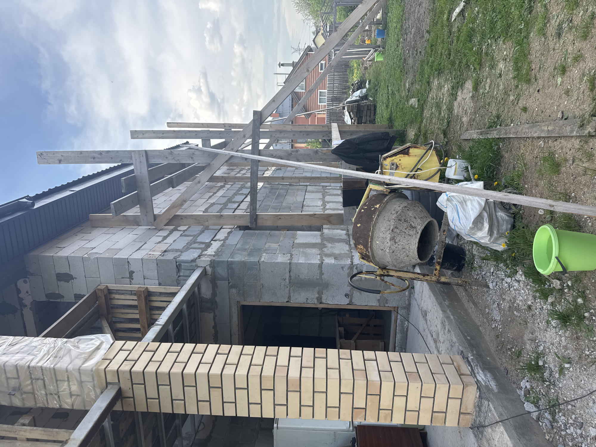 The image shows a construction site in Perm, Russia, where a building is being erected. The structure is made of bricks and cinder blocks, with wooden scaffolding providing support. A cement mixer is present on the site, along with a green bucket, bags of materials, and various construction tools. The environment appears to be a residential area with houses visible in the background. The sky is cloudy.