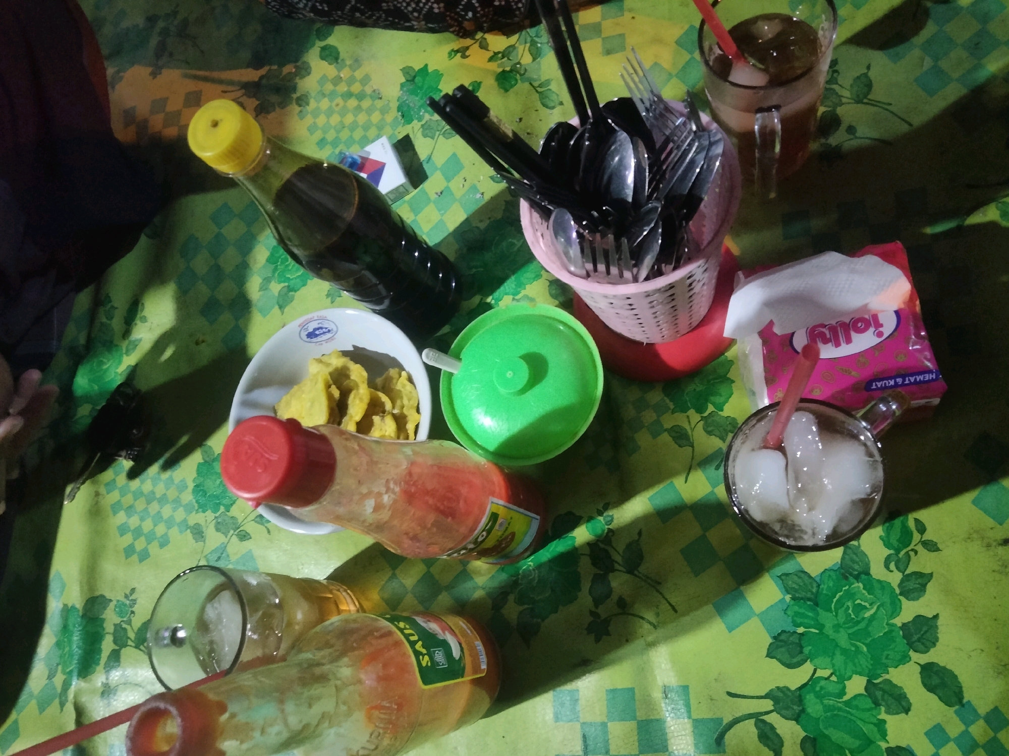 The image depicts a table setting with food and drinks, possibly in Merakurak, Indonesia. A green checkered tablecloth covers the surface. On the table, there is a bottle of dark liquid with a yellow cap, a white bowl containing what appears to be fried food, a green plastic container with a lid, a pink basket holding various utensils like spoons and forks, and two glasses of iced beverages. One glass contains a dark liquid with ice, and a red straw. Another glass is tipped over, showing ice and liquid. There are also two sauce bottles with red caps visible. A package of tissues with the brand name "Jolly" is also present. The lighting suggests an indoor setting, potentially at dusk or night, with some shadows cast on the table.