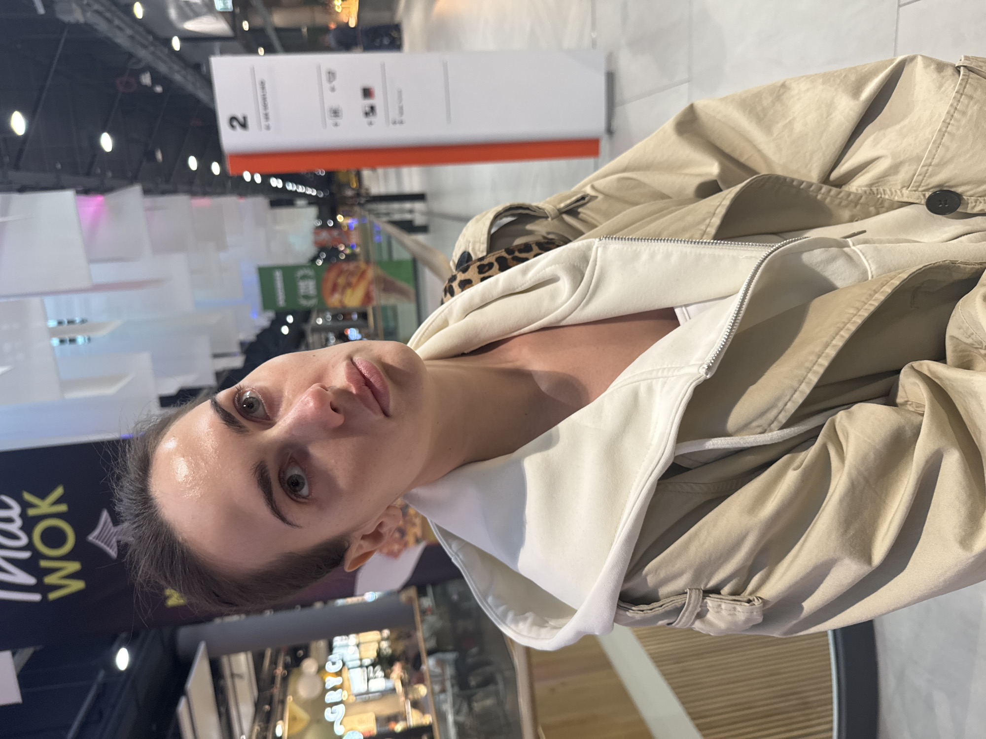 A young woman is captured in a close-up, correctly oriented portrait shot within a brightly lit indoor commercial space, likely a shopping mall or food court in Warsaw, Poland. She looks directly at the camera with a neutral expression. Her dark hair is pulled back, and she wears a light-colored, possibly off-white or cream, zip-up hoodie or jacket, layered under a light beige or khaki trench coat. A leopard print strap is visible over her right shoulder.
The background is bustling with activity, featuring modern architecture and artificial lighting. To her left, a prominent black sign with yellow text reads "MAU WOK," accompanied by a star-like symbol, indicating a food establishment. Further back and to her right, a tall white vertical sign with an orange top stripe displays the large number "2," along with smaller, unreadable text and icons below it, possibly marking a section or entrance. Other blurry signs and commercial displays are visible in the background, reinforcing the setting as a public commercial area. The overall atmosphere suggests daytime or active operating hours.