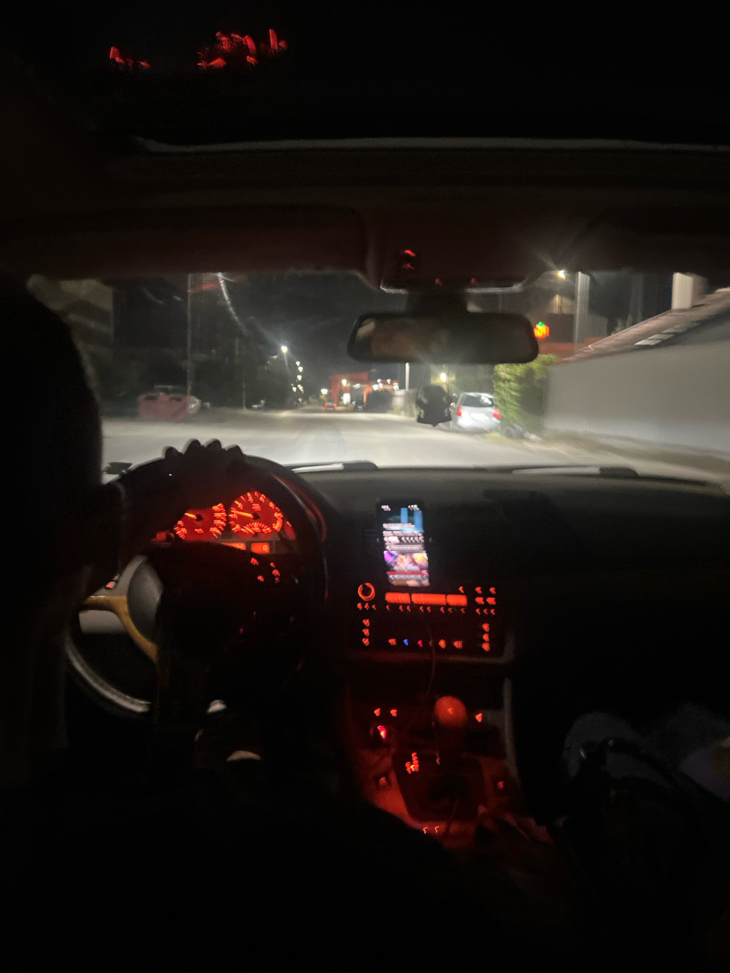 The image captures the interior of a car at night, seen from the passenger's perspective. The dashboard is illuminated with red lights, highlighting the car's instrument panel and central console. A person's shoulder and the back of their head are visible on the left side of the frame, suggesting they are the driver. The car is in motion, as indicated by the blurry streetlights and buildings visible through the windshield. The scene appears to be in a suburban area, with houses and streetlights lining the road. The location is identified as Montana, Bulgaria.
