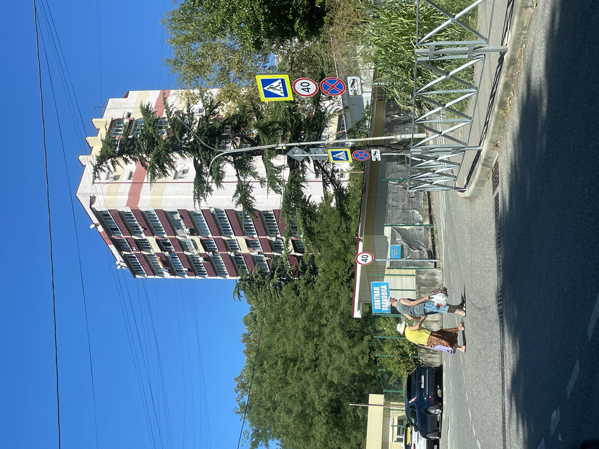 The image shows a sunny day scene on a city street in Sochi, Russia. In the background, a multi-story apartment building with a red and yellow striped facade stands tall against a clear blue sky, with power lines running across it. To the left, lush green trees fill the frame, partially obscuring the lower levels of the building.
In the foreground, two elderly individuals, a man and a woman, are crossing a street at a designated crosswalk. The woman, on the left, is wearing a yellow shirt and a patterned skirt, and is barefoot. The man, on the right, is wearing a grey sleeveless shirt and blue shorts. Both are carrying shopping bags. They appear to be walking together.
Beside the crosswalk, a blue sign with white text reads "ПЛАТНАЯ ПАРКОВКА", which translates to "PAID PARKING". Several traffic signs are mounted on a lamppost to the right of the crosswalk, including pedestrian crossing signs, a speed limit sign indicating 40, and prohibitory signs. A metal fence with a diamond pattern lines the right side of the road. A dark blue car is parked on the left side of the street, and part of a white car is visible next to it. The road surface is asphalt with white lane markings. The environment suggests a residential or mixed-use urban area.