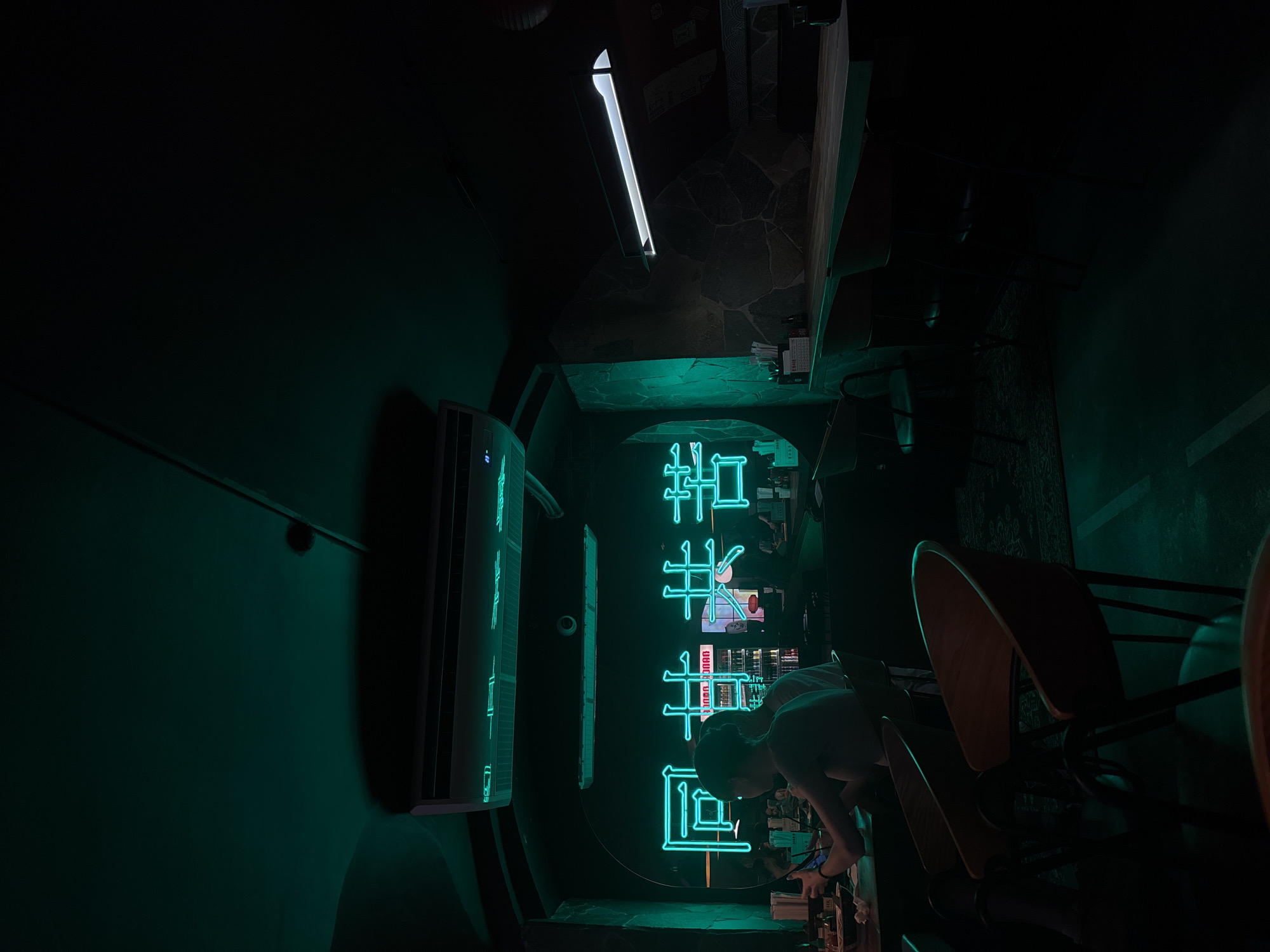 The image shows the interior of a dimly lit space with a strong emphasis on neon lighting. A vertical neon sign in a stylized Asian script is a prominent feature. The setting appears to be an establishment in Perm, Russia. A person is bent over a counter or table, with chairs nearby. An air conditioning unit is mounted on the wall on the left. The scene is tight, suggesting a small or narrow space.