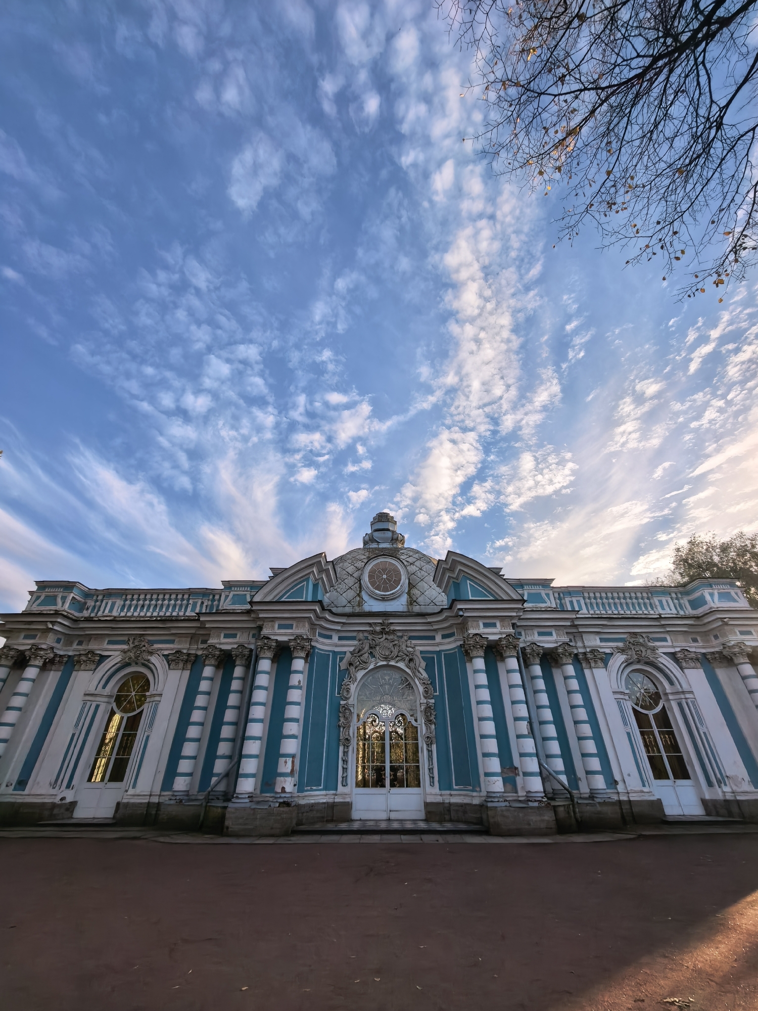 The image shows a low-angle, wide shot of the Hall of Ceres at the Catherine Palace complex in Pushkin, Russia. The building is ornate, featuring turquoise and white stripes, fluted columns, and arched windows. A large, decorative clock is visible at the top center of the facade. Above the building, a bright blue sky is filled with wispy white clouds, and the bare branches of a tree are silhouetted against the sky on the right side of the frame. The ground in front of the building is a reddish-brown dirt path, with some fallen leaves. The sun is low in the sky, casting a warm light on the scene.