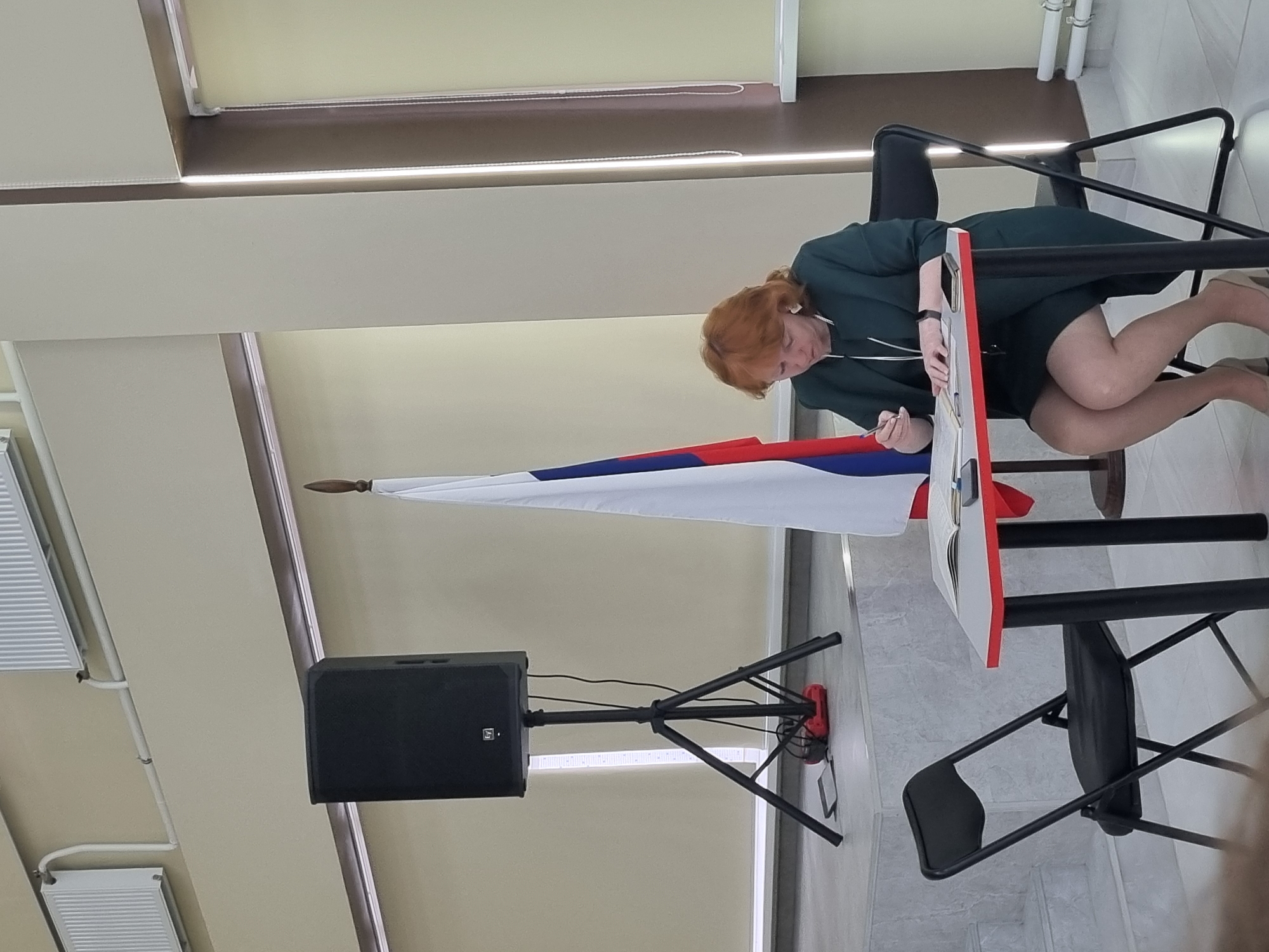 A woman with red hair is seated at a table, writing in a notebook. She is wearing a dark green dress and beige heels. A Russian flag stands behind her. To her left is a black speaker on a tripod stand. To her right is a black folding chair. The setting appears to be an indoor event space, possibly a classroom or auditorium, with blinds on the windows and radiators on the wall. The lighting is ambient and diffused. The specific location is the city of Chaikovsky, Russia.