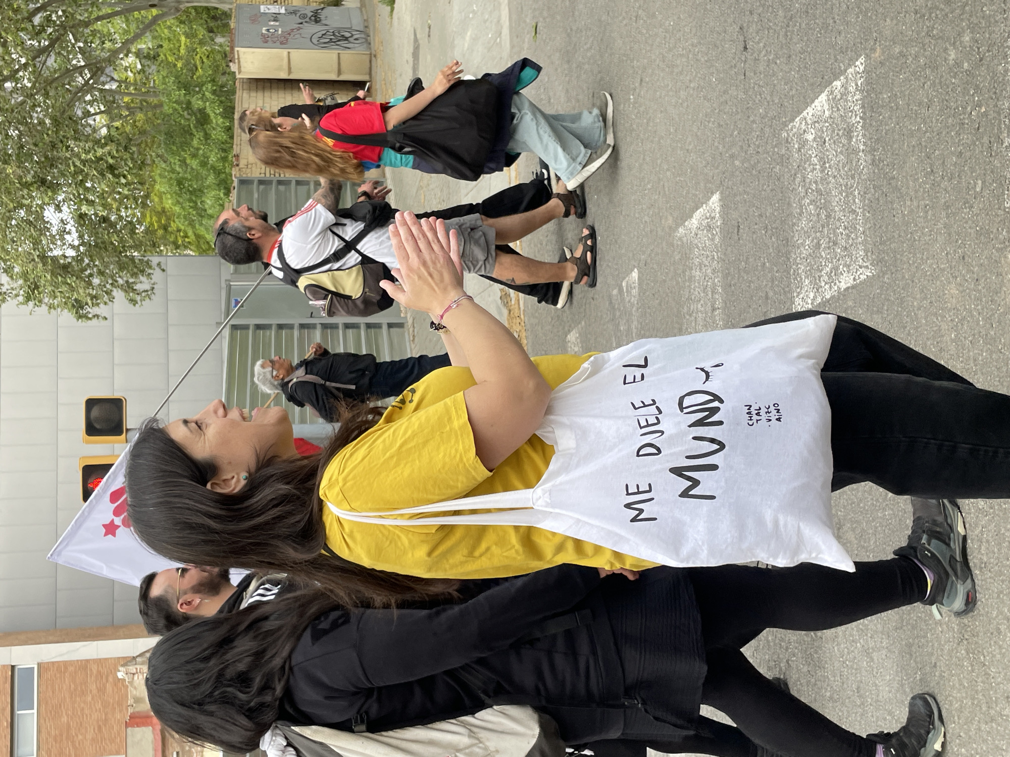 The image captures a vibrant street scene in Barcelona, Spain, under bright daylight, suggesting a sunny day with distinct shadows on the pavement. People are walking along an urban street, possibly participating in a procession or public gathering.
In the foreground, a woman with long dark hair, wearing a bright yellow t-shirt and black pants, is captured mid-stride. She carries a white tote bag over her shoulder with the text "ME DVELE EL MUND" (likely a stylized "ME DUELE EL MUNDO" - "The world hurts me") and smaller words "CHAN TAL . VIZC AINO" printed on it. Her face is turned upwards, eyes closed, with a wide smile or laugh, indicating joy or excitement. A small turquoise earring is visible in her left ear. To her left, another person in black attire is partially visible.
Behind her, in the midground, a man with a dark beard and short dark hair is seen. He wears a white t-shirt with red accents, grey shorts, and dark sandals, carrying a backpack. His hands are raised in a gesture that appears to be clapping or waving. Further back, another woman with long light brown hair is walking, dressed in a red t-shirt, light blue jeans, and carrying a black tote bag. An older individual with grey hair is visible further back on the right.
The background features typical urban elements: a building with graffiti, green trees, and a traffic light showing a red signal. A white flag with a red star and crescent is partially visible on the left side of the frame, held by an unseen individual. The overall atmosphere appears lively and positive, with participants engaged in the event.