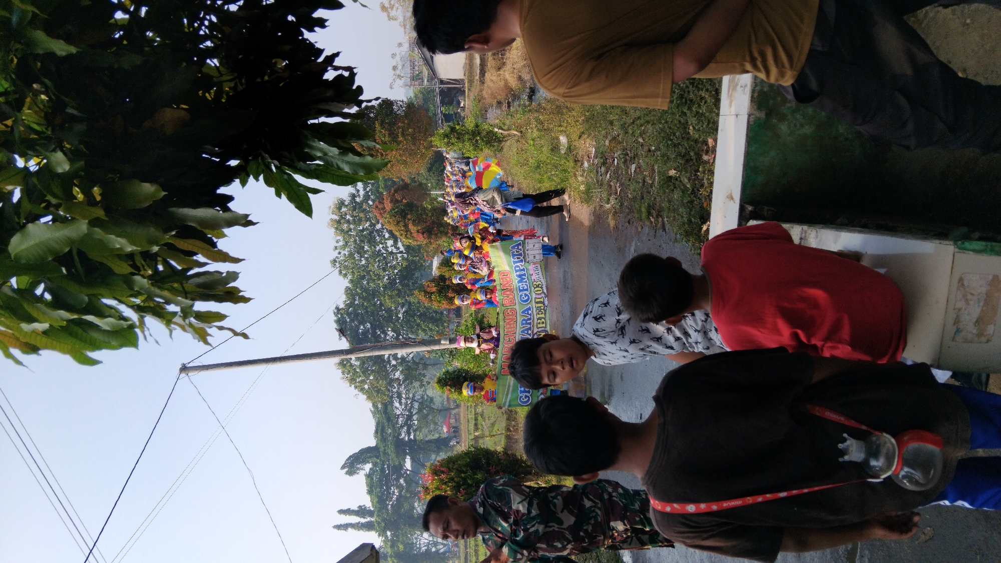 In Posong, Indonesia, a street parade is in progress, featuring a marching band from the 'GITA SUARA GEMPITA' organization, identified by a banner they carry. The participants, mostly children dressed in vibrant uniforms and hats, are marching down a paved road lined with trees and vegetation. A man in military attire stands on the left, observing the parade, while several children and a man in a tan shirt are positioned in the foreground, watching the procession. The environment appears rural, with fields visible in the background and a narrow waterway running alongside the road. The sky is clear and bright, suggesting it is daytime.