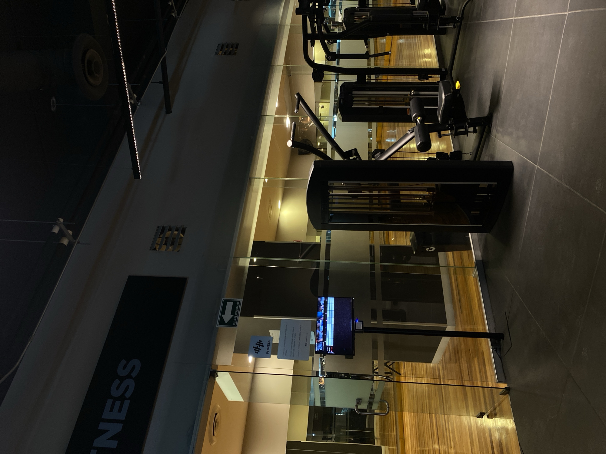 The image depicts a dimly lit interior space, seemingly a gym or fitness center, in an unidentified city. The floor is covered in gray tiles. In the foreground, various black weight machines are visible. The back wall is a transparent glass partition, revealing a wooden floor in the adjacent room, and various equipment inside the gym. A television screen is mounted on a pole near the glass door. Near the door, signage indicating the name of the gym (partially visible as "FITNESS") along with an evacuation route sign are attached to the glass.