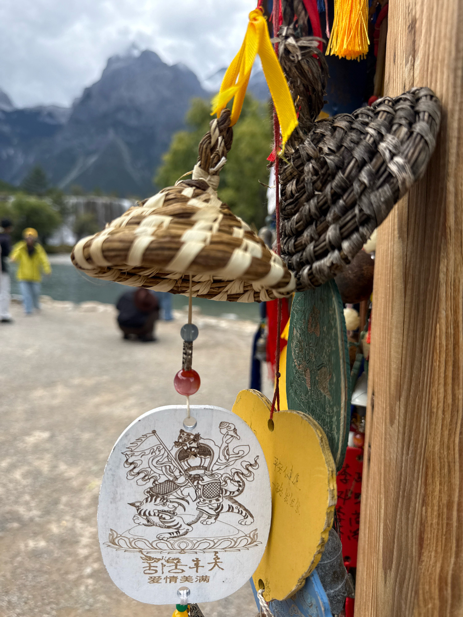 In Yulong Nakhi Autonomous County, China, a close-up shot focuses on a collection of hanging charms attached to a wooden post. The charms include a small woven hat-like object with a braided cord, a heart-shaped yellow charm with faint Chinese calligraphy, and an oval-shaped white charm featuring an intricate line drawing of a warrior riding a tiger, also with Chinese characters below. Beads in red and blue are visible, strung between the charms. In the blurred background, a body of water, a pathway, and distant mountains under a cloudy sky can be seen. Several people are present in the background, some walking and one crouching.