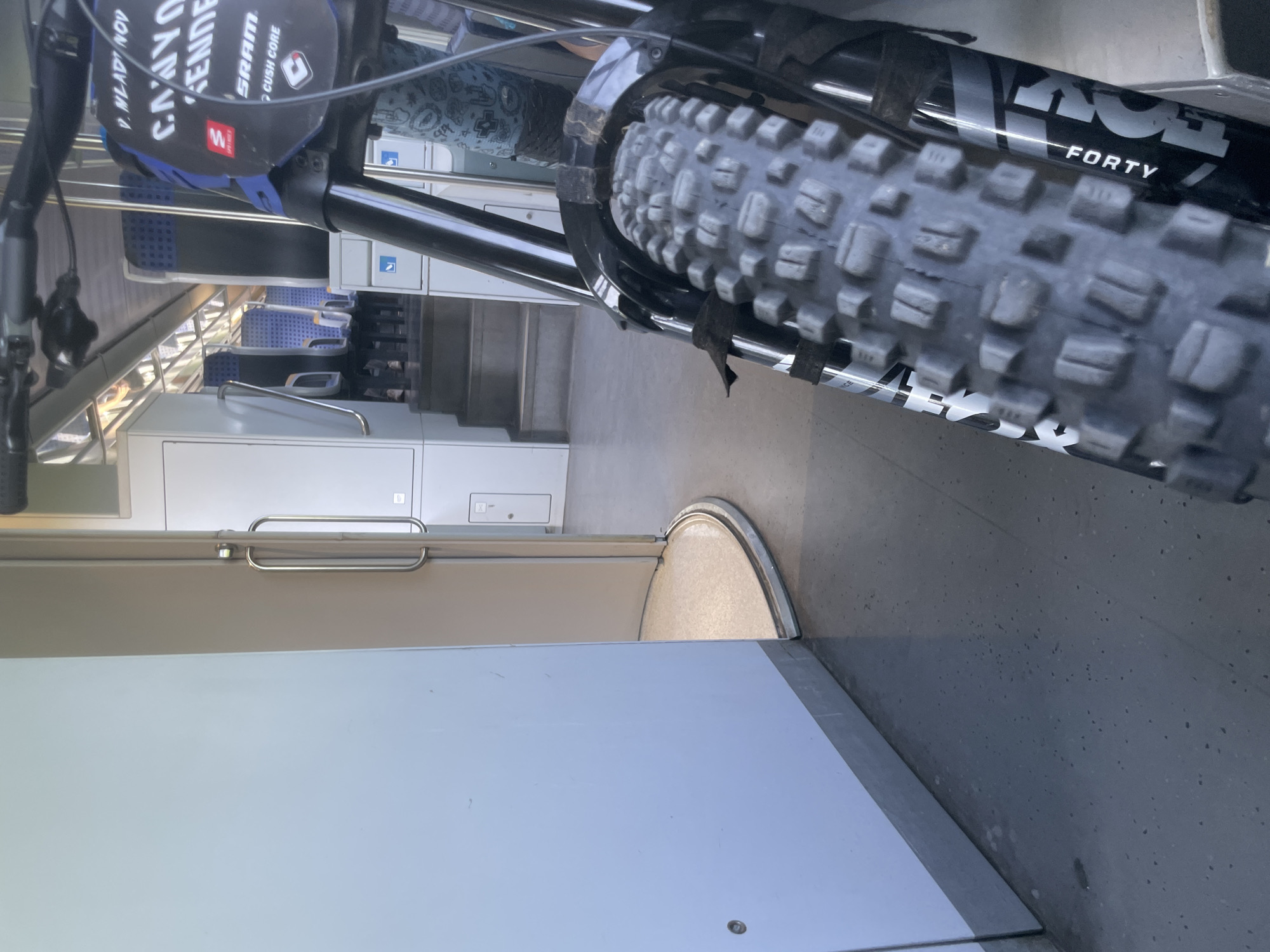The image depicts the interior of a train carriage, possibly in Merichleri, Bulgaria, though this location is not explicitly visible. A mountain bike is positioned in the foreground, taking up a significant portion of the right side of the frame. The bike's front wheel and suspension fork are prominently featured, with the "FOX 40" branding visible on the fork. The background shows rows of empty blue and gray train seats, suggesting the train is either at a station or during a quiet period of travel. The lighting is artificial, typical of an indoor train environment. To the left of the bike, a white wall dominates the space, with a partially open door revealing a curved walkway and a glimpse of the train's exterior or another section of the interior. The floor is a dark, speckled material. There are no people clearly visible in the image. The overall impression is one of travel and possibly the anticipation of an outdoor activity, given the presence of the mountain bike.