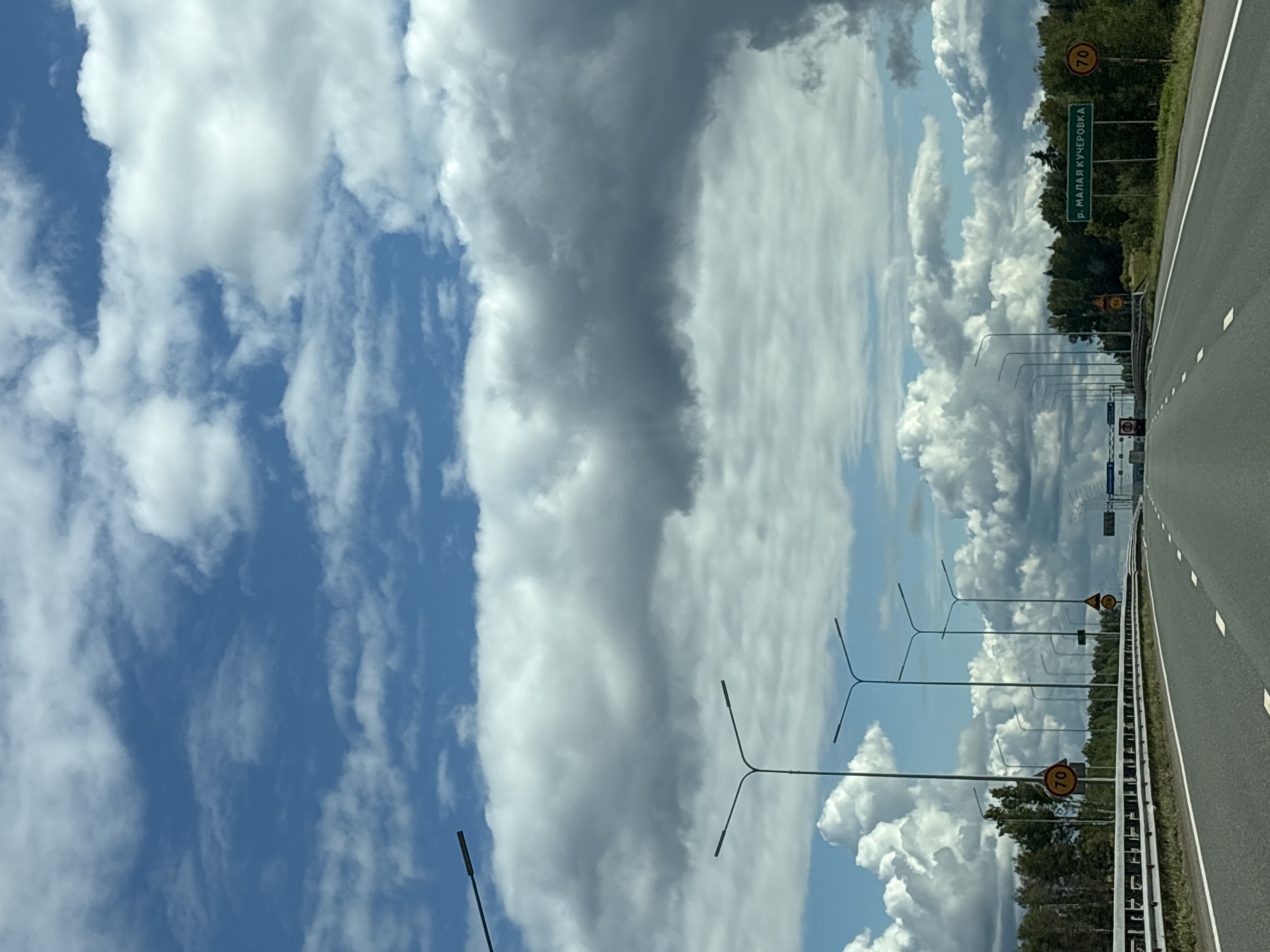 The image shows a highway scene under a partially cloudy blue sky. The perspective is from the driver's seat of a vehicle, looking forward. On the right side of the highway, there are several streetlights and trees. A green road sign with white lettering reads "р. МАЛАЯ КУЧЕРОВКА", indicating a place name. There are also two circular speed limit signs, one showing "70" and another partially visible sign below it. On the left side of the highway, there is a metal guardrail and more trees in the distance. Several more streetlights are visible on the left side as well. The road itself is asphalt with white lane markings. In the far distance, there appears to be a toll booth or border crossing with some vehicles. The overall environment suggests a rural or semi-rural highway setting. The location is identified as Zvyozdny, Russia.