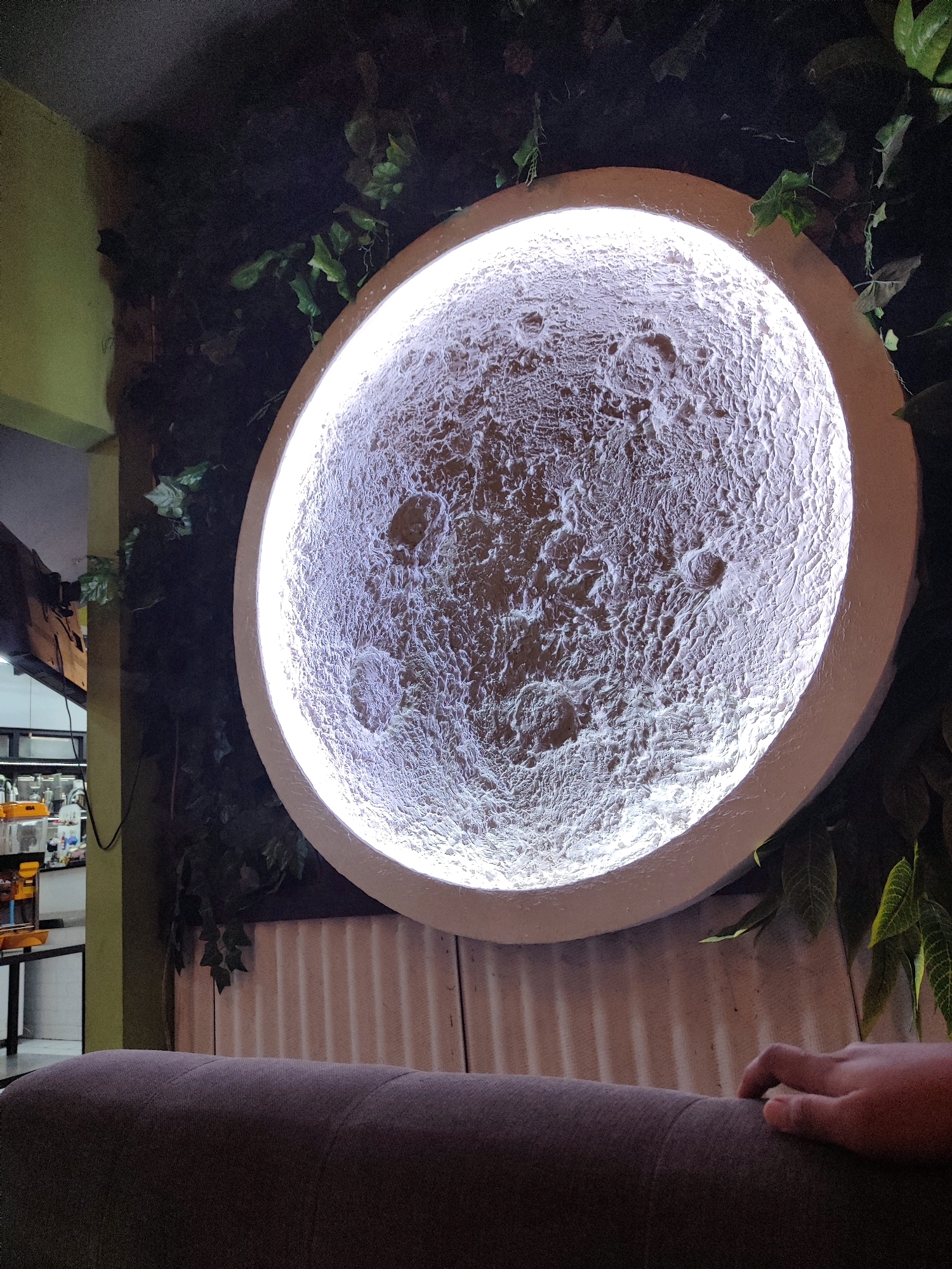 An illuminated, textured circular object resembling the moon is mounted on a dark, leafy wall. The object is lit from behind, creating a soft glow. In the foreground, a portion of a dark grey sofa is visible, with a person's right hand resting on its surface. To the left of the moon-like object, a yellow wall and a section of shelving with various items are partially visible. The overall scene appears to be indoors, possibly in a cafe or lounge area in Blindungan, Indonesia.