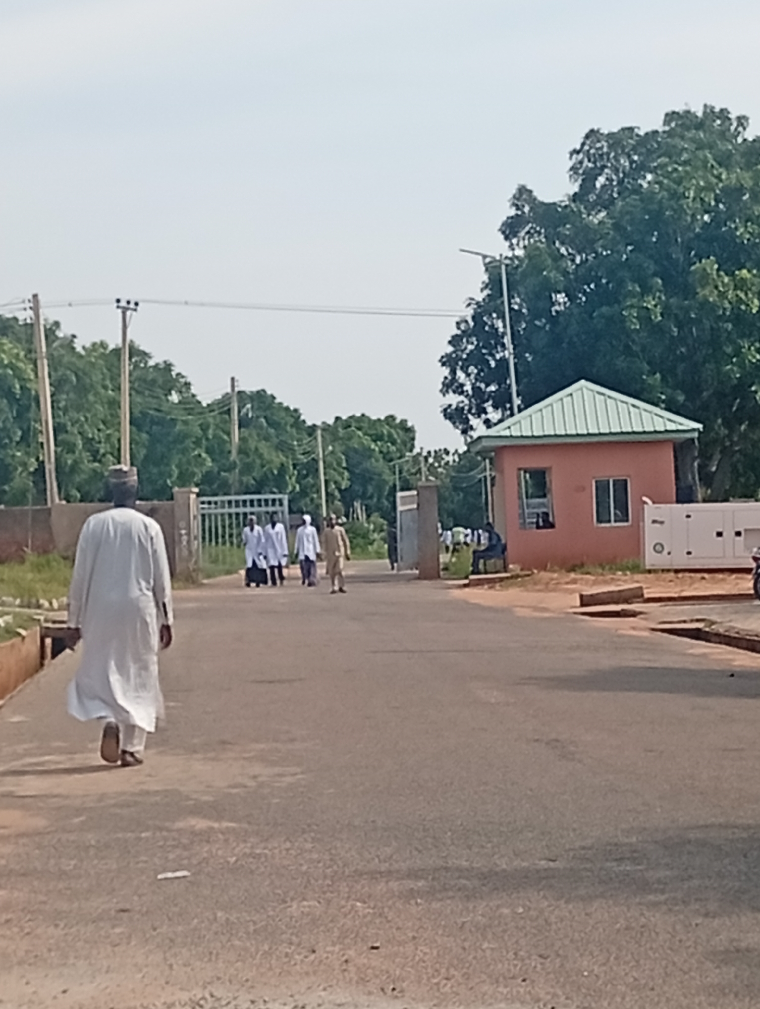 The image depicts an outdoor scene, likely within the grounds of a facility in Gombe, Nigeria. In the foreground, a person dressed in a white flowing robe and a cap walks away from the camera on a paved road. Several other individuals, also in white robes, are seen walking together further down the road towards a gate. To the right of the road, a small building with a green roof stands, possibly a guardhouse or a small office. A large white generator or industrial unit is situated next to this building. The environment is characterized by trees and vegetation, with utility poles and power lines visible in the background. The sky is overcast. The scene appears to be a daytime shot, possibly a normal day on the premises.