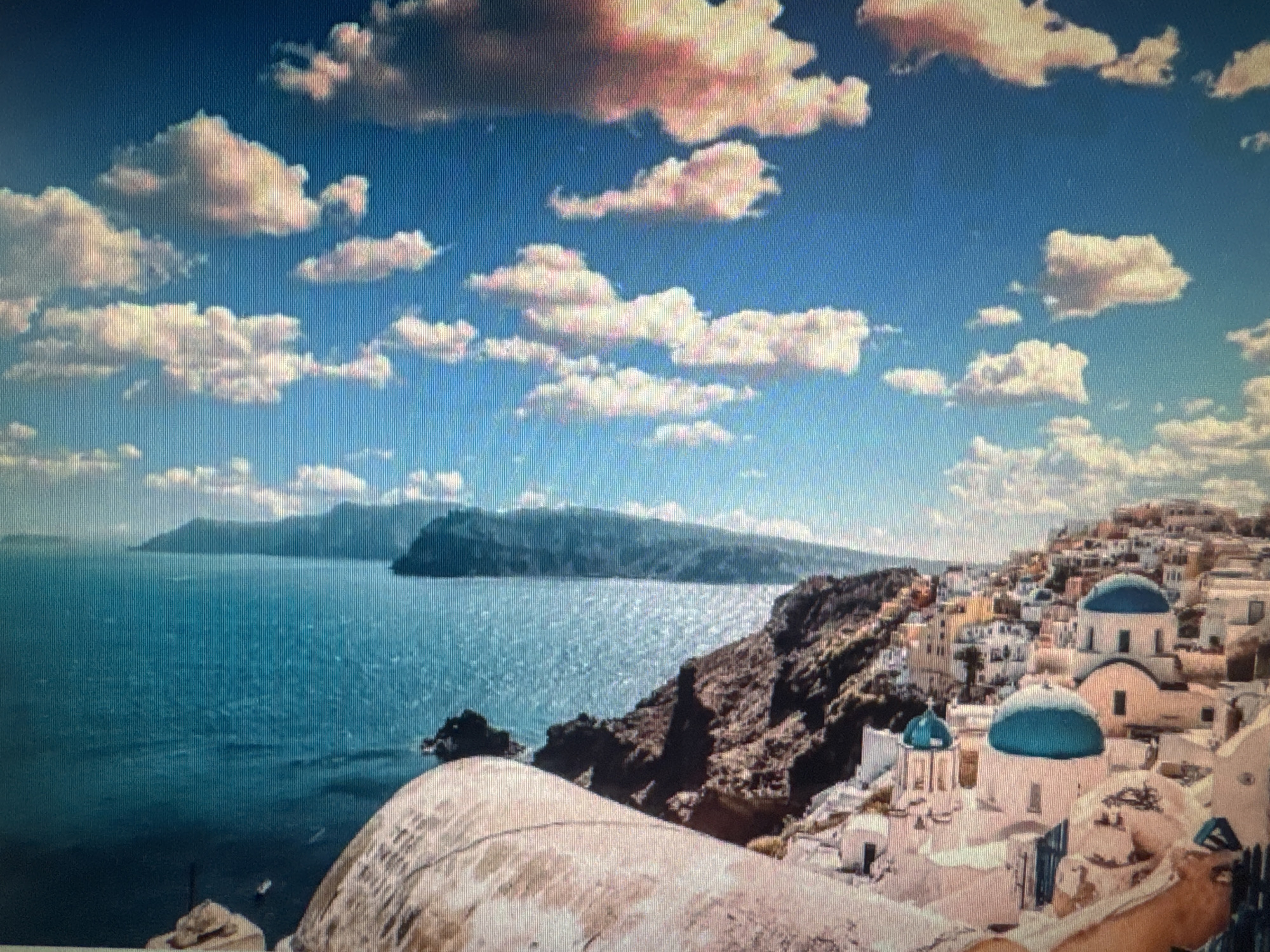 This media file depicts a picturesque coastal scene with a village nestled on a hillside overlooking the sea. The foreground features a large, textured architectural element, possibly a dome or a rounded building, with a rough, light-colored surface. Beyond this, a vibrant blue sea stretches out towards a distant, hazy coastline with rolling hills.
The village itself is a prominent feature, characterized by whitewashed buildings with distinctive blue-domed roofs. The architecture is clustered tightly on the steep incline, creating a charming and iconic Mediterranean or Aegean aesthetic. Small pathways and steps wind through the village.
The sky above is a brilliant blue, adorned with fluffy white clouds, some tinged with pink and orange, suggesting either early morning or late afternoon light. The sun appears to be bright, casting a warm glow on the scene and creating highlights on the water's surface. The sea is calm, with gentle ripples reflecting the sunlight. The overall mood is tranquil and idyllic, conveying a sense of peace and beauty.
While no people are clearly visible, the presence of boats on the water and the inhabited nature of the village suggest human activity. The location, with its characteristic architecture and coastal setting, strongly resembles Santorini, Greece, a popular tourist destination known for its white villages and blue domes. There is no visible text in the image. The image does not contain any elements that specifically relate to Ellwangen, Germany.