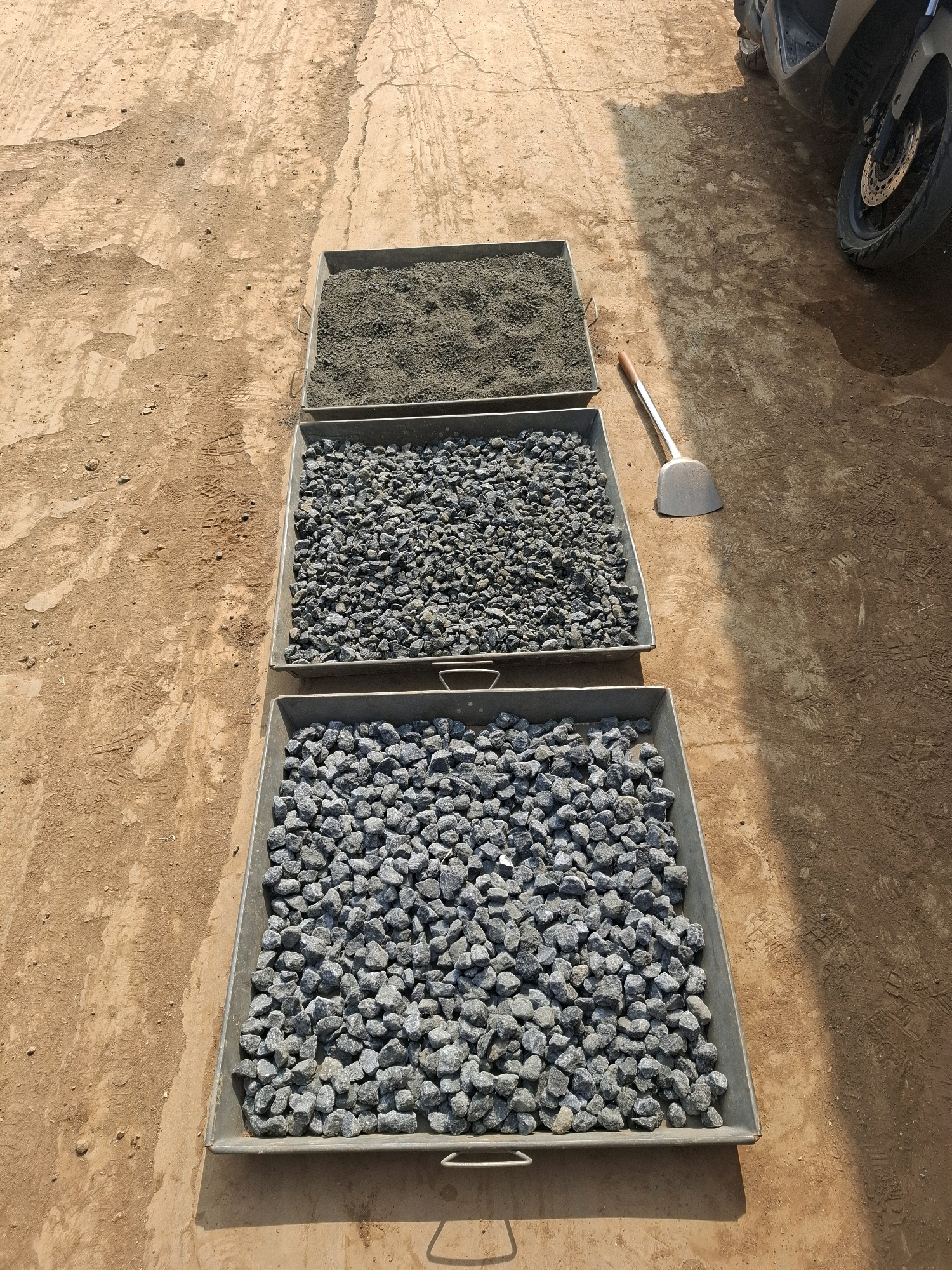 Three metal trays are arranged in a row on a dirt surface. The tray furthest from the viewer contains fine, dark gray material, possibly soil or sand. The middle tray is filled with medium-sized gray gravel. The tray closest to the viewer contains larger gray gravel. A metal shovel with a wooden handle rests on the ground between the top two trays. In the background, a portion of a parked scooter or motorcycle is visible, its rear wheel and part of its body in shadow. The ground appears to be a mixture of packed dirt and sand with tire tracks and footprints imprinted. The scene is outdoors, under sunlight, casting shadows. The location is identified as Ciheulang Tonggoh, Indonesia.