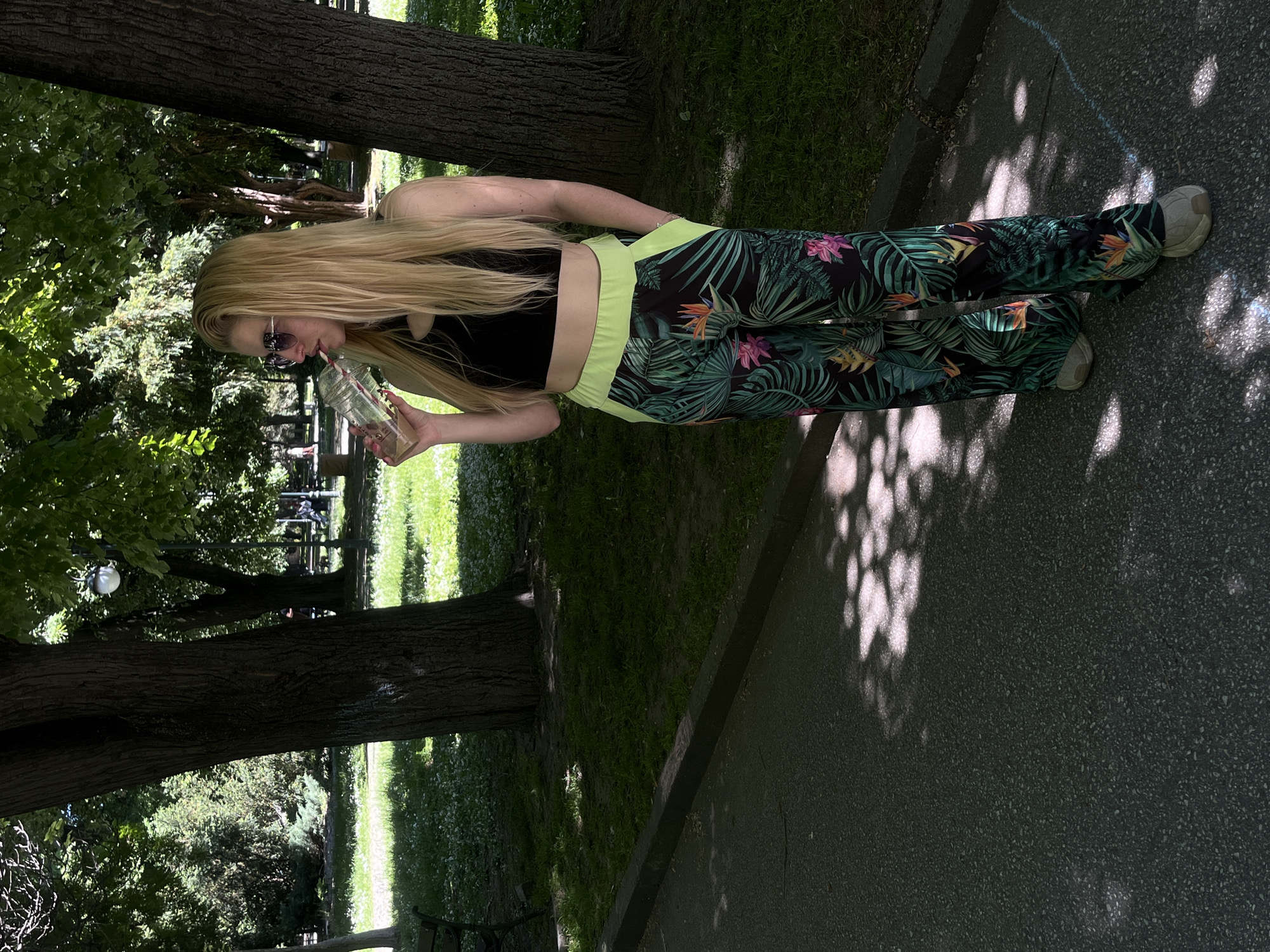 A woman stands in a park on a sunny day, drinking from a plastic cup. She wears a black crop top, tropical print pants with neon green waistband, and sneakers. She has long blonde hair and sunglasses. The park has trees, grass, and a paved pathway with shadows. The location appears to be a park in Plovdiv, Bulgaria, based on the scene's characteristics.