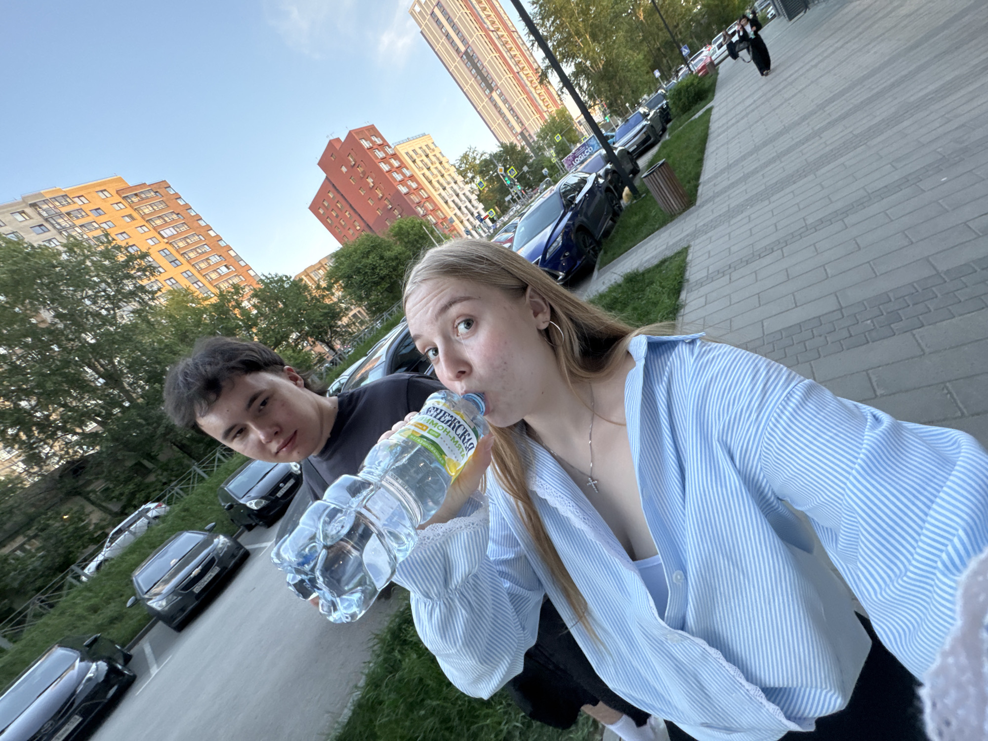 A young woman with blonde hair is drinking from a plastic water bottle labeled "СНЕЖСКАЯ ЛИМОН-МЯТА". She wears a blue and white striped shirt and a silver necklace with a cross pendant. A young man with dark hair is standing beside her, looking at the camera. They are outdoors on a paved area next to a grassy strip where several cars are parked. In the background are apartment buildings of varying colors and heights, as well as a walking person in a black dress. The scene is in Perm, Russia.