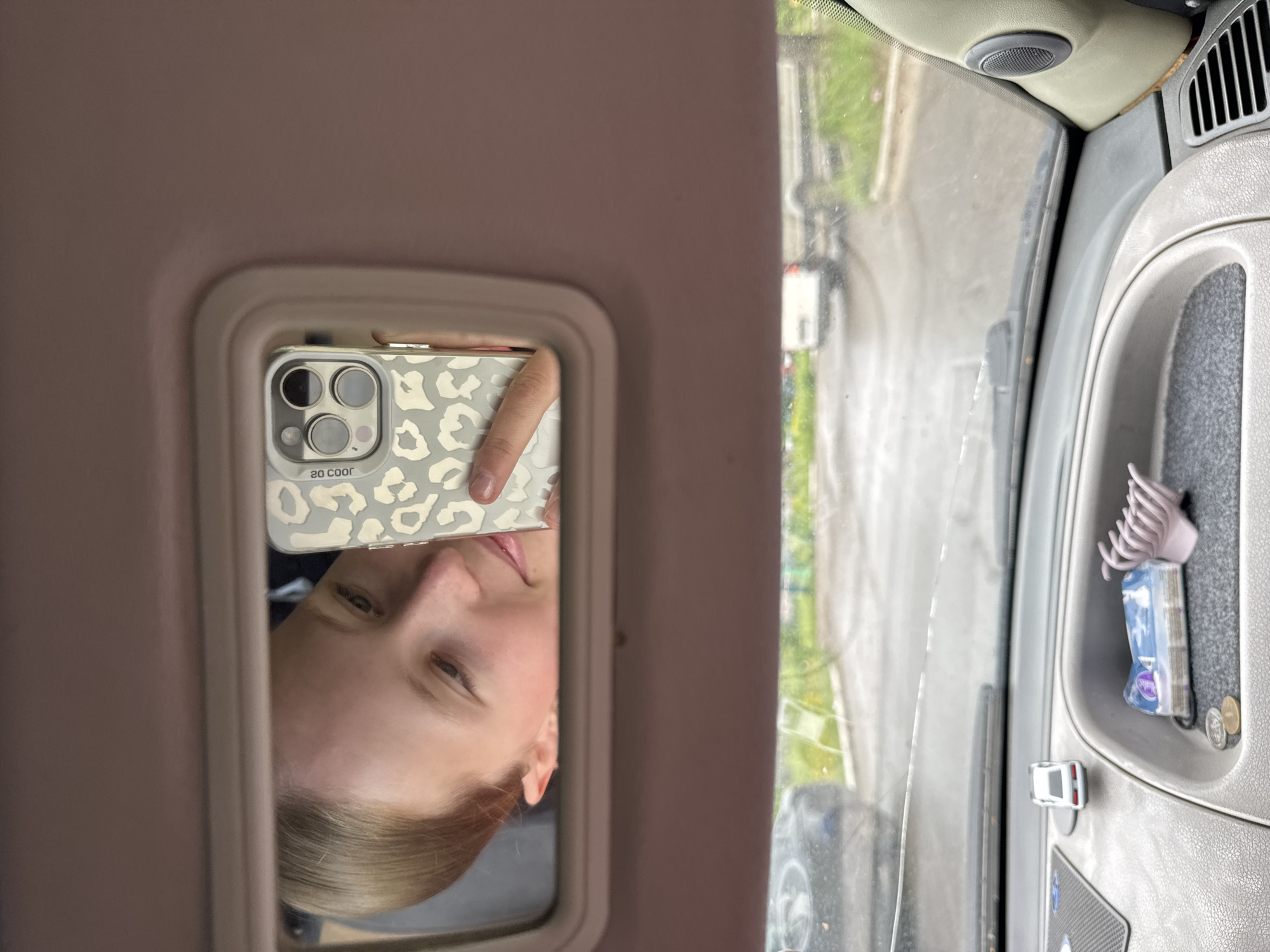 The image displays a candid selfie captured by a young woman's reflection in a car's vanity mirror. The woman, possessing blonde hair and a fair complexion, shows a calm demeanor with a slight, subtle smile. She is holding a smartphone enclosed in a light grey and white leopard print case, on which the text "20 coor" is visible. Her hand, with painted nails, is partially seen gripping the phone.
The rectangular mirror is set within a light-colored panel of the car's interior, likely a sun visor. To the right of the mirror, the vehicle's interior features grey plastic surfaces, including a speaker grill and air vents located near the windshield. Below these elements, a recessed storage area contains a lavender claw hair clip, a pack of tissues, and two coins. A small white toy car is also visible on an adjacent ledge.
Through the vehicle's windshield, a blurred daytime view of a street or road is visible, appearing wet or reflective, which might suggest recent rain. Green foliage and indistinct background structures hint at an urban or suburban setting. The scene is set inside a vehicle, presumably in Chaikovsky, Russia. The image documents the act of taking a self-portrait within this everyday environment.