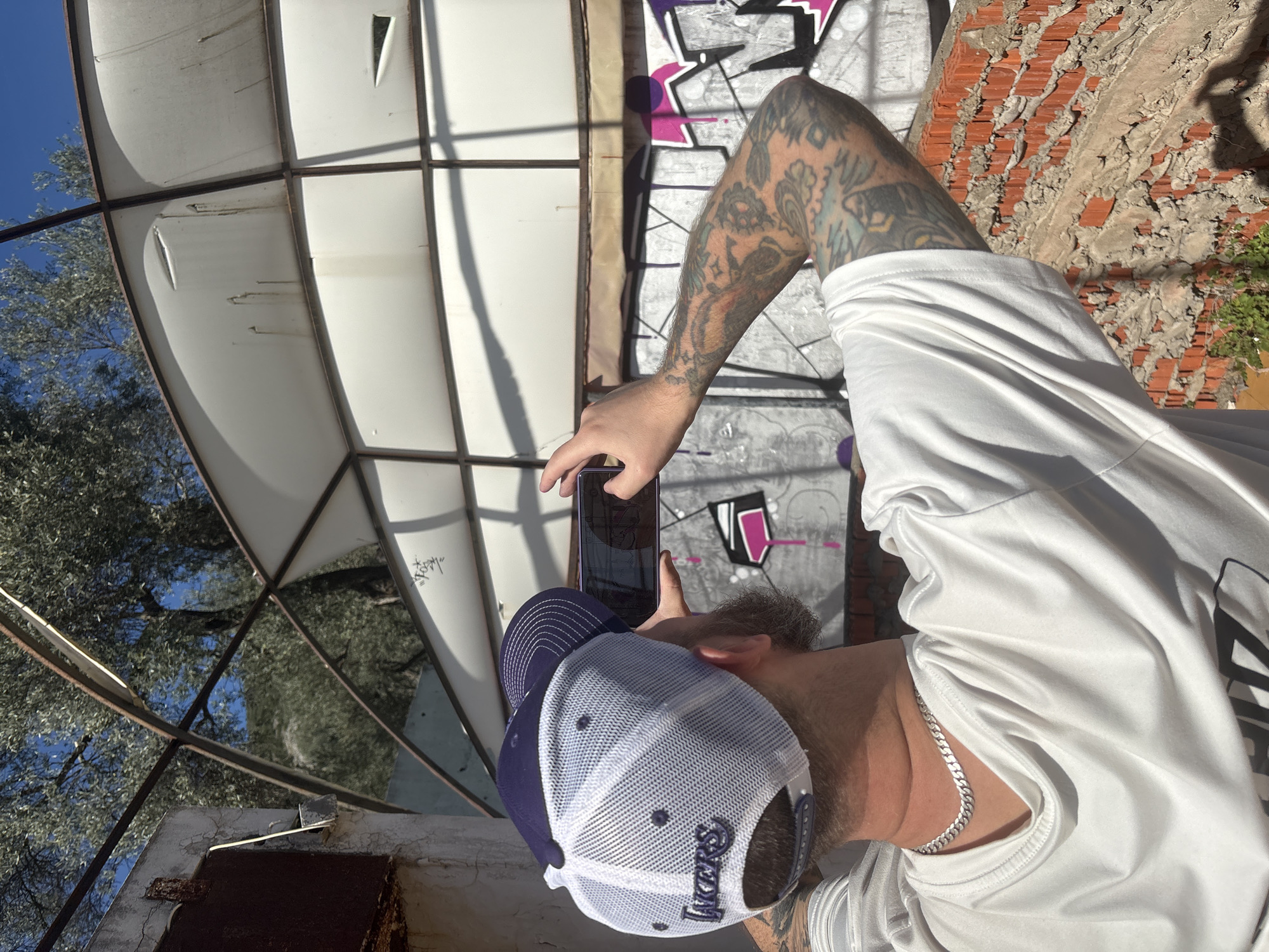 A person wearing a white t-shirt, silver chain, and a blue and white Lakers baseball cap is holding a smartphone to take a photo. The person has a tattooed arm. The setting appears to be an abandoned structure with graffiti on the walls. The structure has a metal frame and some transparent paneling that provides a view of trees and the sky. Brickwork is also visible. The photo is taken in Bratica - Braticë, Montenegro.