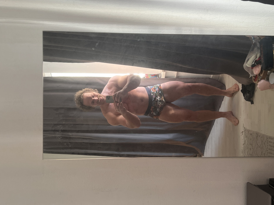The image displays a full-body mirror selfie of an adult male captured indoors. The man, of Caucasian appearance, has light brown curly hair and a beard, and exhibits a muscular physique. He is shirtless and wears patterned boxer briefs. He is smiling and holds a light-colored smartphone (consistent with an iPhone) with both hands at chest level, positioning it to photograph his reflection. He stands barefoot on a light-colored floor, which features a partially visible small, light-colored rug on the left side of the frame. The background reflected in the mirror includes dark gray curtains, which partially obscure a bright light source, likely a window, on the right. Miscellaneous items are visible on the floor to the right, including a black bag, some light-colored fabric or clothing, and a pink, possibly ceramic, object. The wall visible outside the mirror frame on the left is plain white. The specific location within the city of Saint Petersburg, Russia, cannot be determined from the visual content of the image.