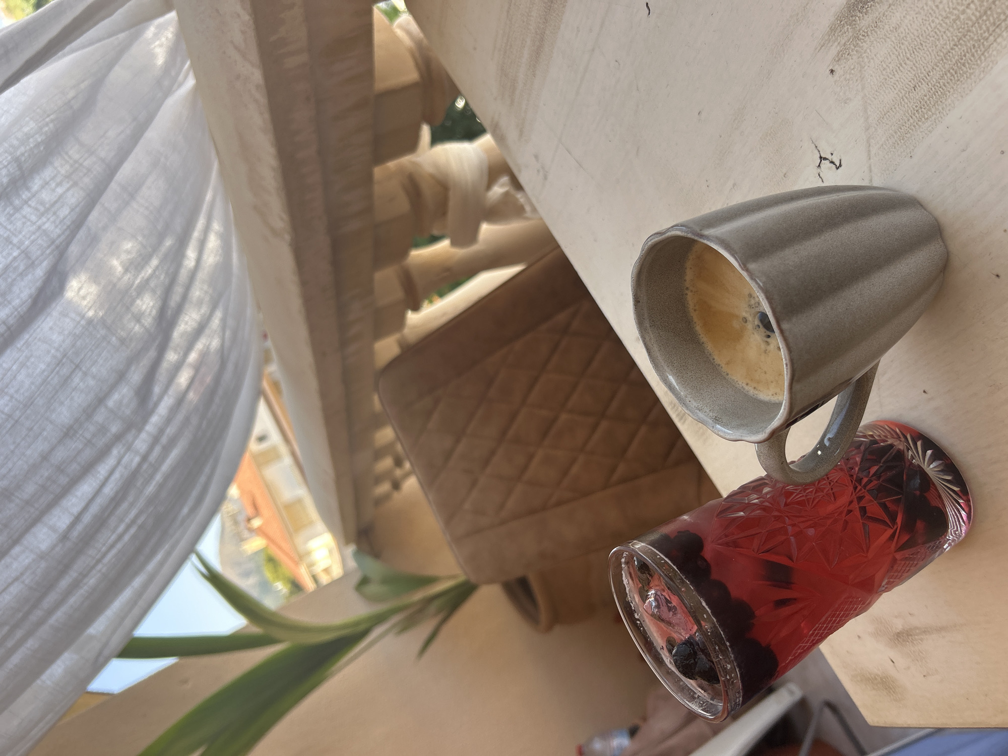 The image captures a close-up of a table setting on an outdoor patio, likely in Capdepera, Spain, as suggested by the distant view of terracotta-roofed buildings and hills under a bright sky. A tall, patterned glass filled with a red beverage and dark berries rests on the left side of the frame. To its right sits a ribbed ceramic mug containing a dark coffee with crema. In the background, a quilted upholstered chair is visible, and beyond that, a balcony railing with ornate balusters overlooks the Mediterranean landscape. A sheer white curtain billows gently above the scene, filtering the sunlight. The table itself appears to be made of light-colored, weathered wood. There are no people visible in the frame.